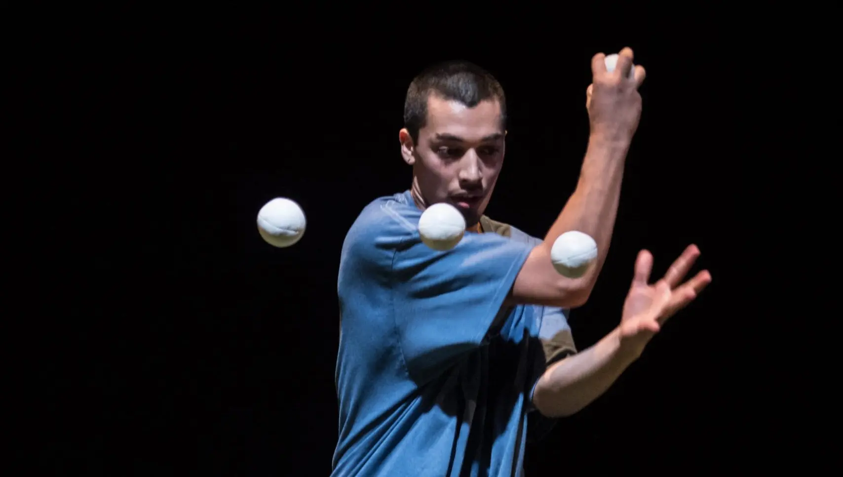 Sereno Aguilar Izzo, en chemise bleue, jongle avec trois balles blanches sur un fond sombre, les bras levés et l'expression concentrée.