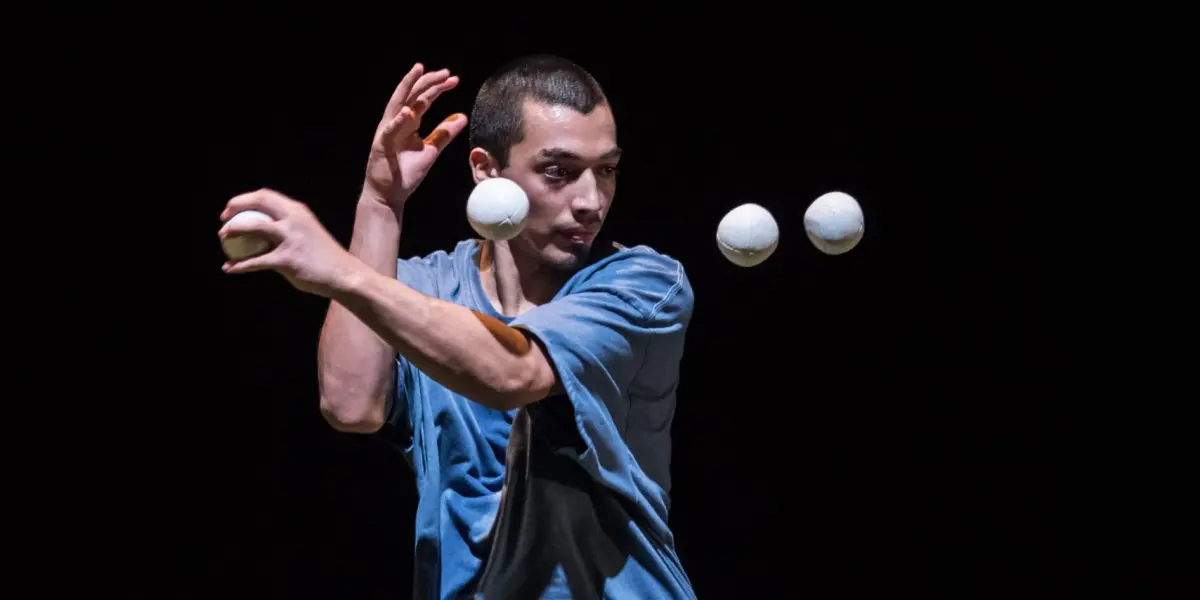 Sereno Aguilar Izzo, en chemise bleue, se concentre intensément tout en jonglant avec quatre balles blanches en l'air sur un fond sombre.