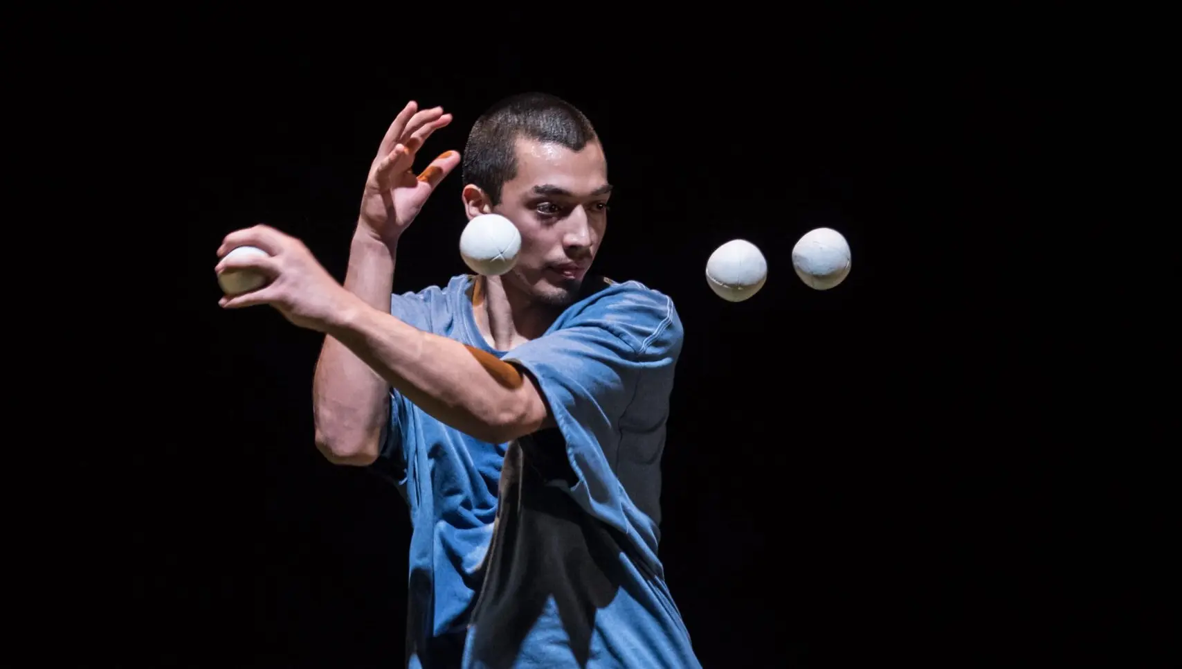 Sereno Aguilar Izzo, en chemise bleue, se concentre intensément tout en jonglant avec quatre balles blanches en l'air sur un fond sombre.