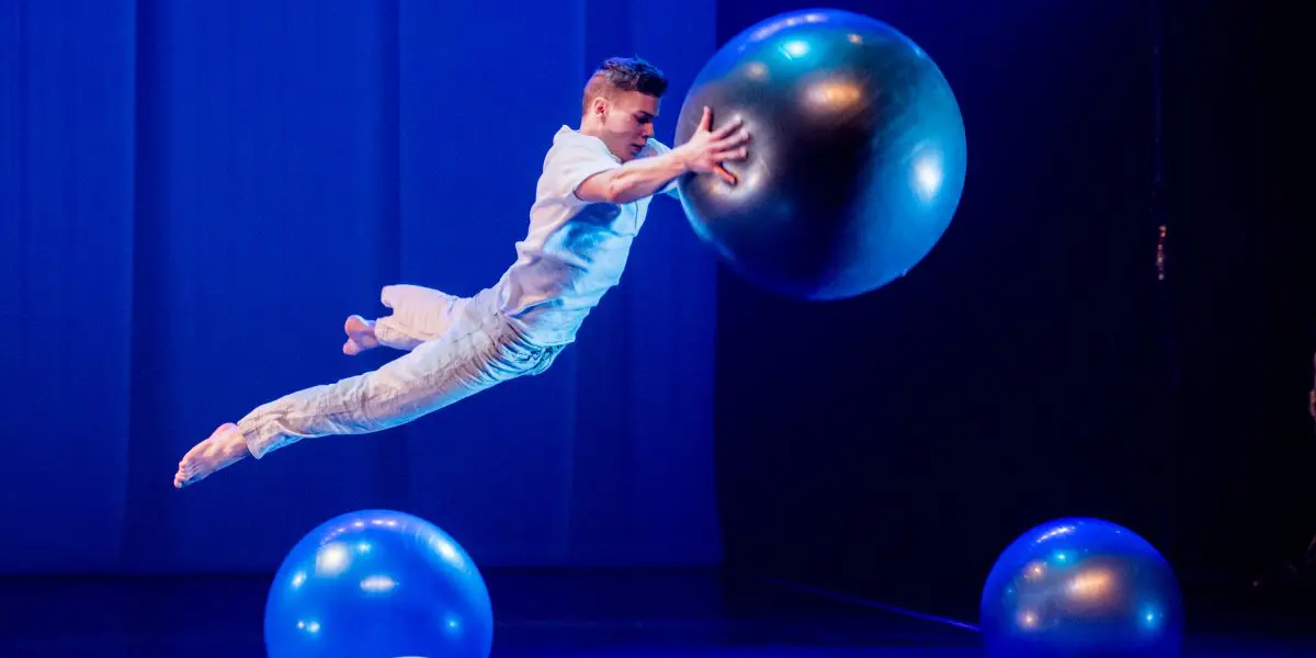 Un interprète en blanc, rappelant le style dynamique de Guillaume Larouche, saute horizontalement en l'air vers une grosse boule bleue sur une scène faiblement éclairée par des lumières bleues et trois autres grosses boules.