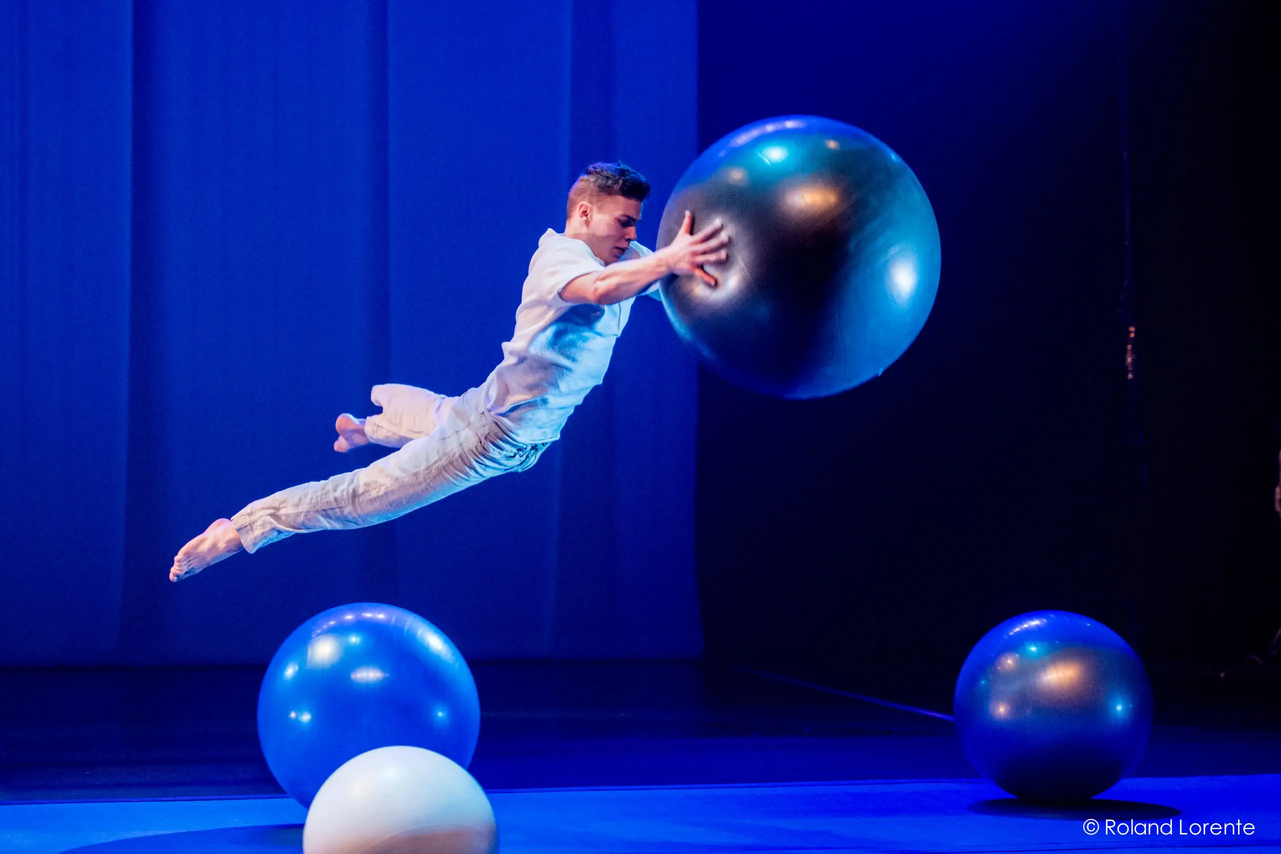 Un interprète en blanc, rappelant le style dynamique de Guillaume Larouche, saute horizontalement en l'air vers une grosse boule bleue sur une scène faiblement éclairée par des lumières bleues et trois autres grosses boules.