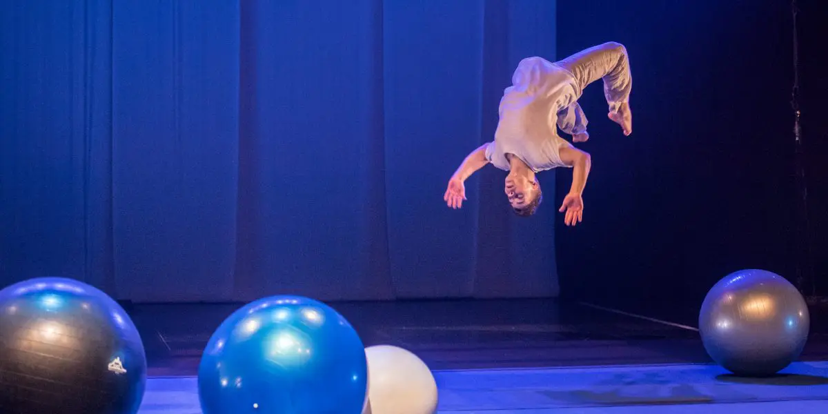 Le performeur Guillaume Larouche, vêtu de vêtements légers, exécute un saut périlleux arrière en plein vol sur la scène, entouré de quatre grands ballons d'exercice colorés, sous un éclairage bleu-violet.