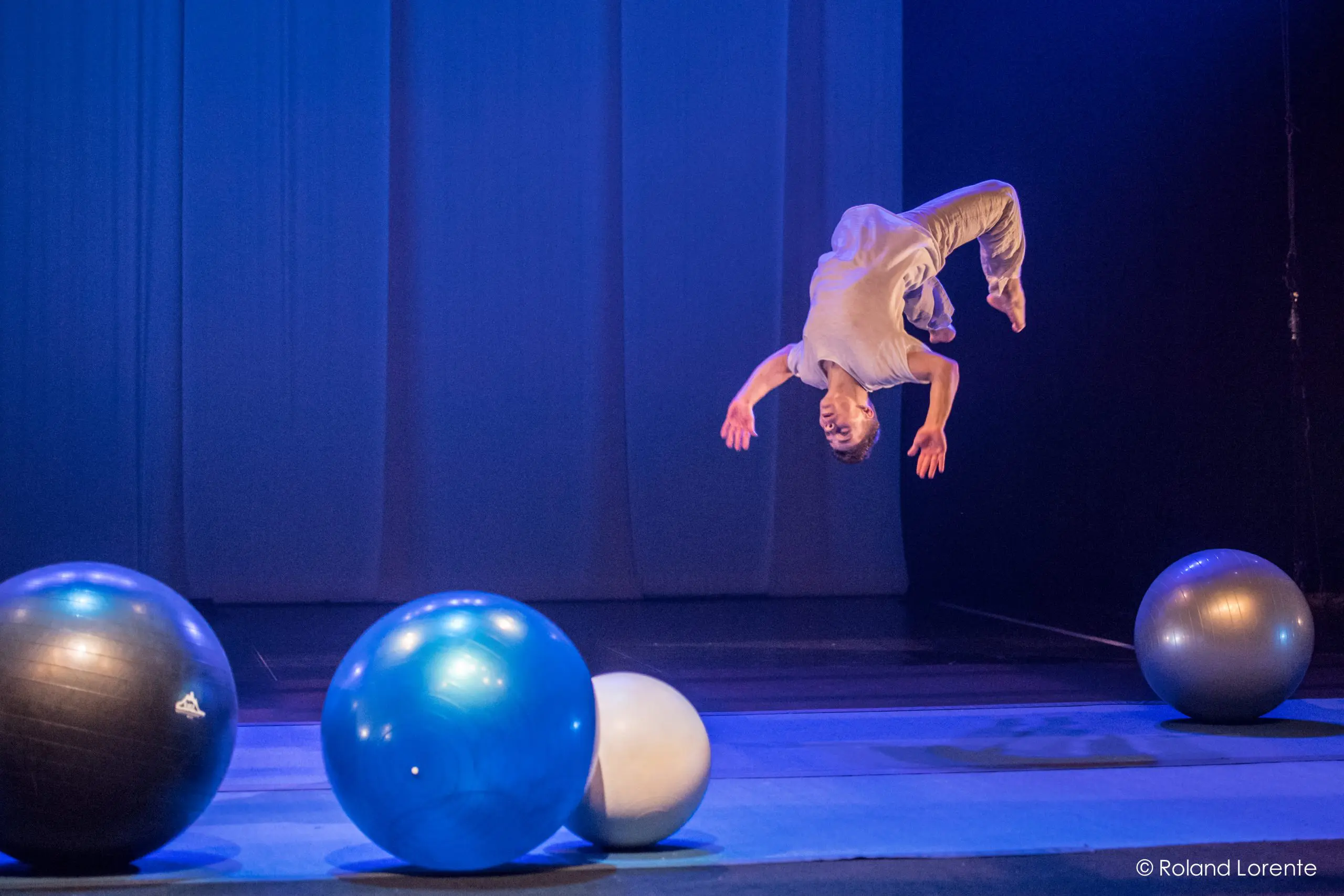 Le performeur Guillaume Larouche, vêtu de vêtements légers, exécute un saut périlleux arrière en plein vol sur la scène, entouré de quatre grands ballons d'exercice colorés, sous un éclairage bleu-violet.