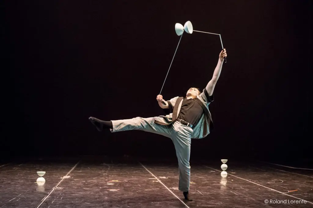 Félix Martin, vêtu d'un costume clair, se tient en équilibre sur une jambe tout en faisant tourner habilement un diabolo sur une corde, d'autres diabolos étant posés sur la scène, sous les projecteurs, sur un fond sombre.