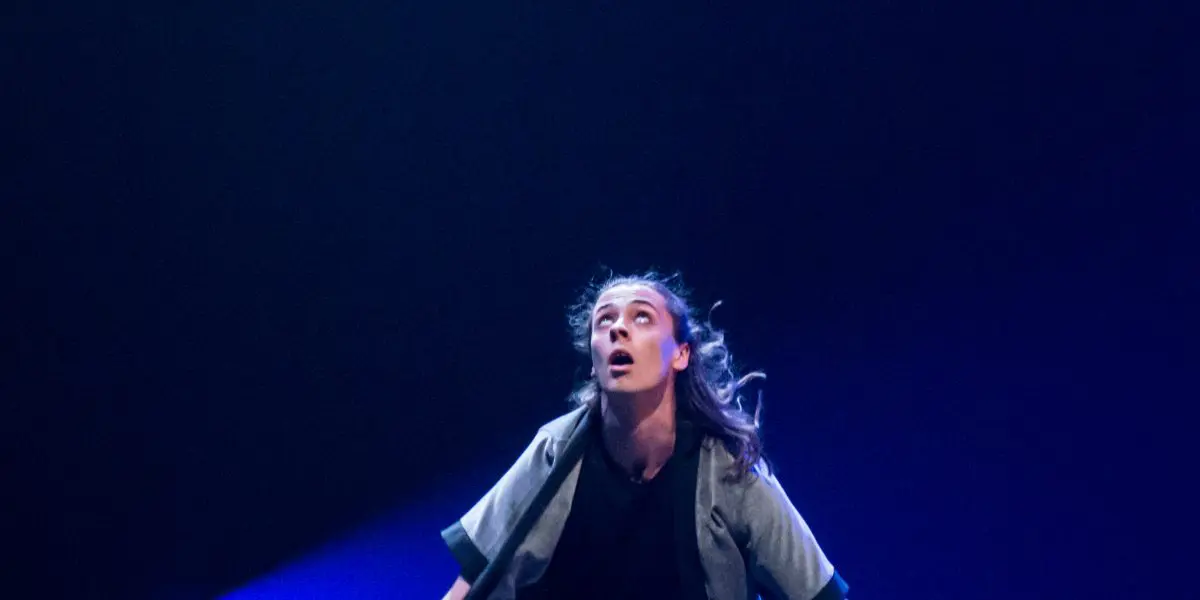 Félix Martin, vêtu de gris, manipule habilement trois yo-yos diabolos sur scène, accroupi et concentré sous un éclairage bleu dramatique.