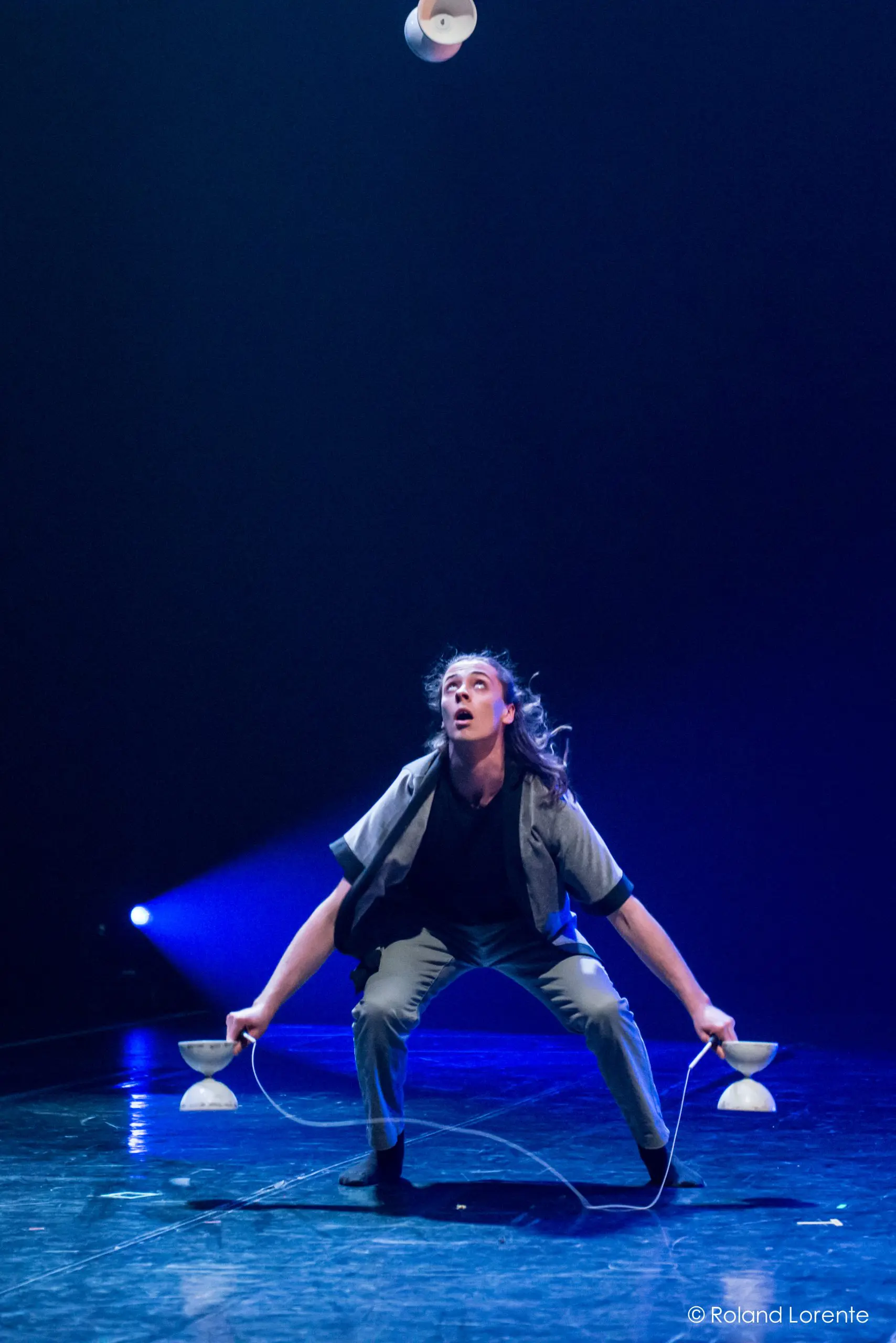 Félix Martin, vêtu de gris, manipule habilement trois yo-yos diabolos sur scène, accroupi et concentré sous un éclairage bleu dramatique.