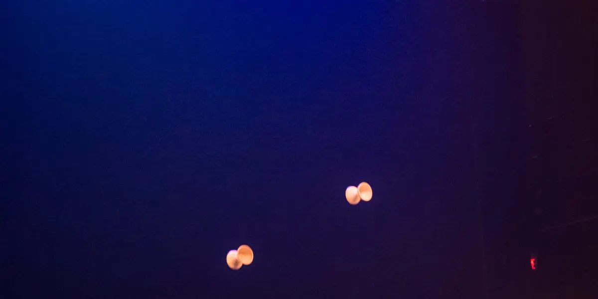 Félix Martin se tient sur une scène sombre sous des lumières bleues, jonglant habilement avec quatre balles blanches munies de ficelles, réalisant un numéro de diabolo envoûtant.