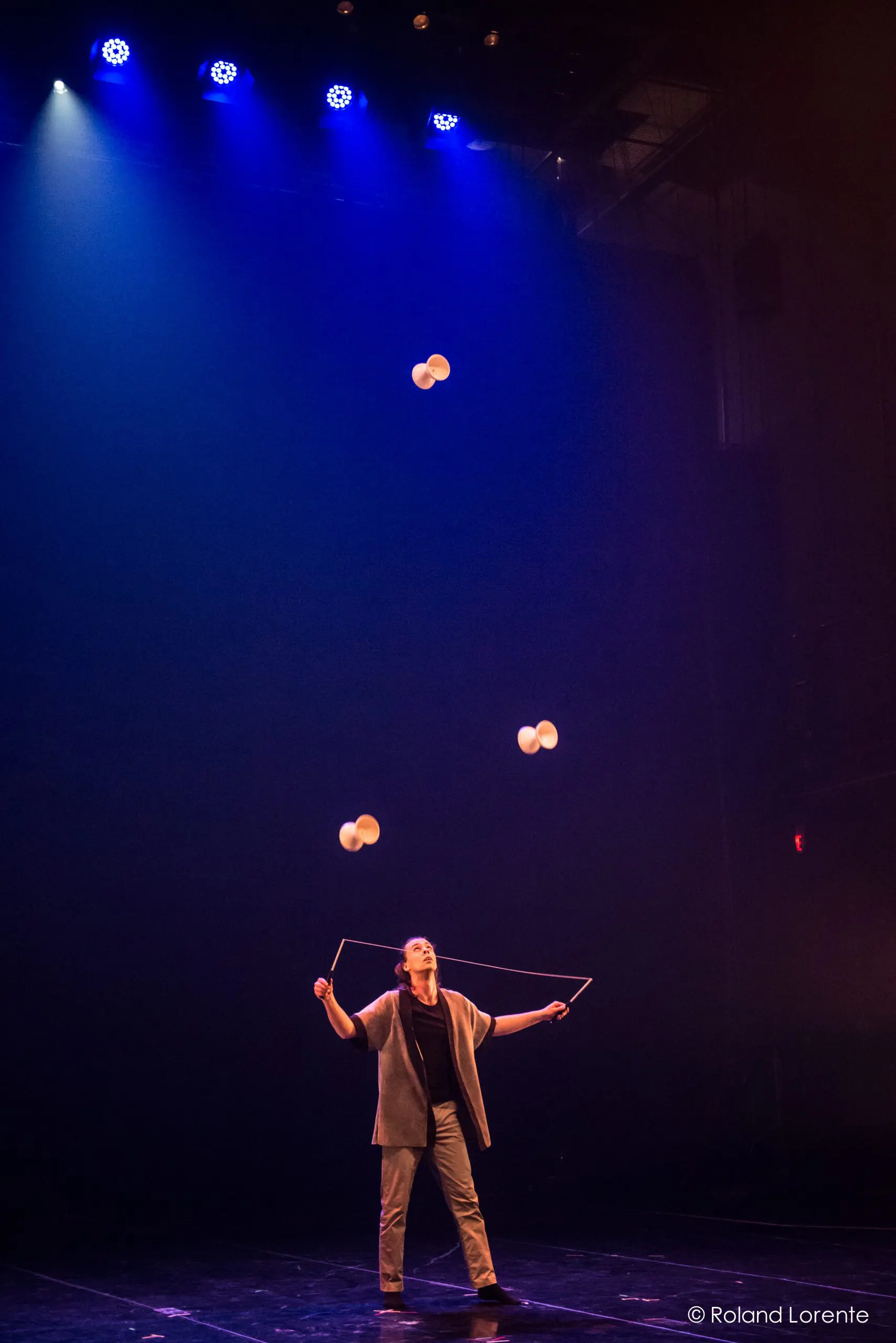 Félix Martin se tient sur une scène sombre sous des lumières bleues, jonglant habilement avec quatre balles blanches munies de ficelles, réalisant un numéro de diabolo envoûtant.
