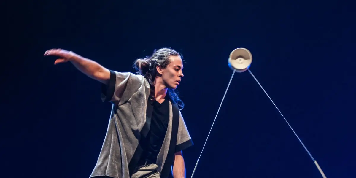 Félix Martin, un artiste sur scène, fait habilement tourner un diabolo avec deux bâtons, vêtu d'un pantalon gris et d'une veste à manches courtes assortie. L'arrière-plan sombre et l'éclairage dramatique mettent en valeur son numéro captivant.