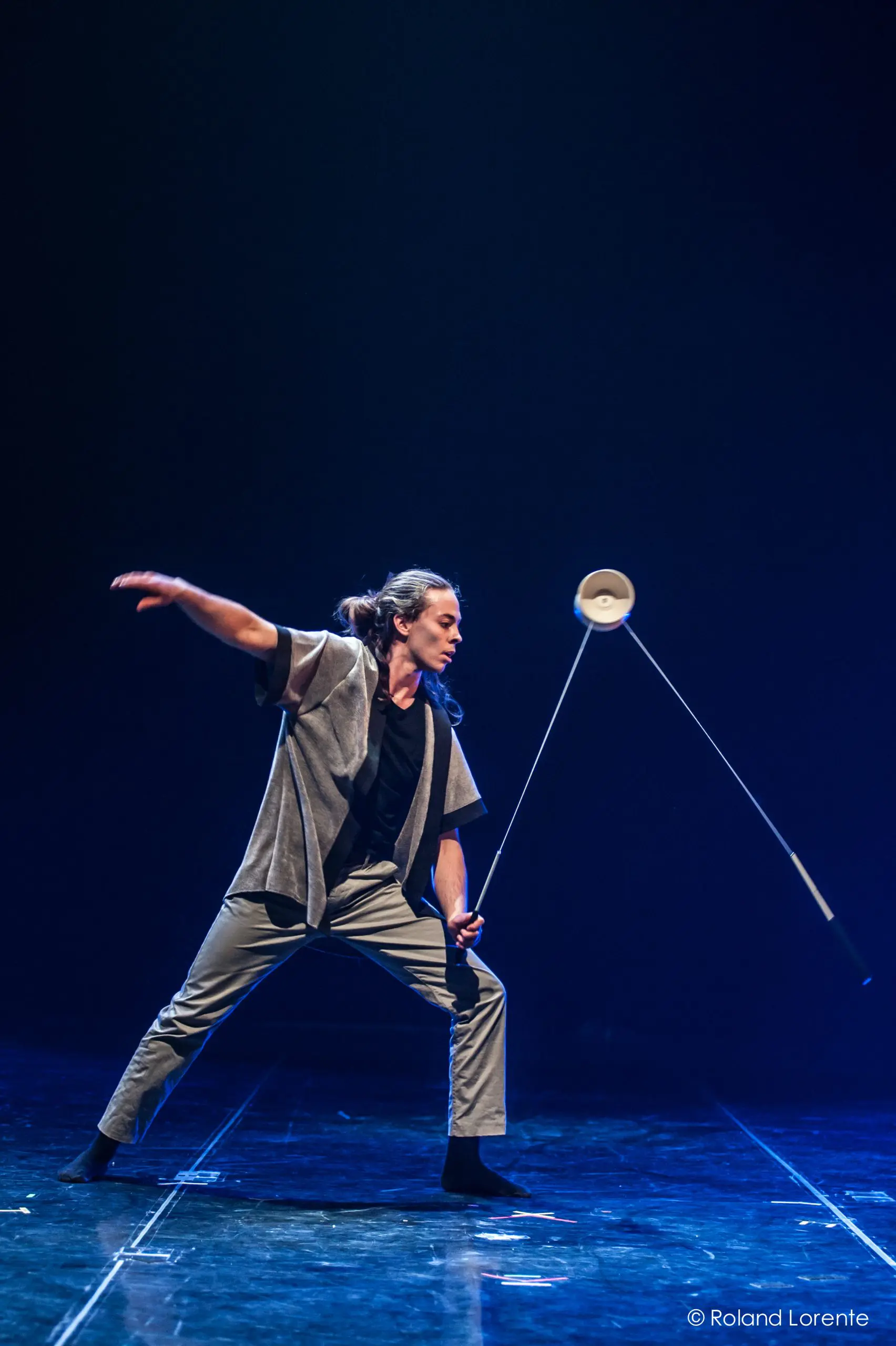 Félix Martin, un artiste sur scène, fait habilement tourner un diabolo avec deux bâtons, vêtu d'un pantalon gris et d'une veste à manches courtes assortie. L'arrière-plan sombre et l'éclairage dramatique mettent en valeur son numéro captivant.