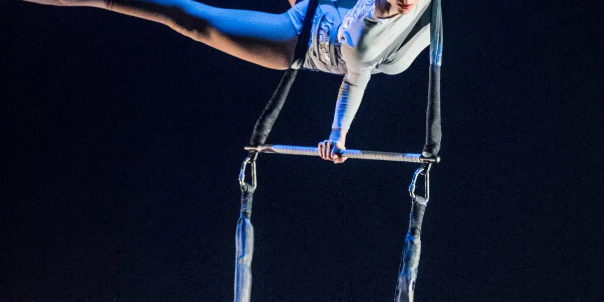 Une artiste aérienne, Halle Baart, vêtue d'un costume blanc, exécute une pose acrobatique sur un appareil suspendu ressemblant à un trapèze, se tenant en équilibre sur un bras tout en étendant gracieusement ses jambes sur un fond sombre.