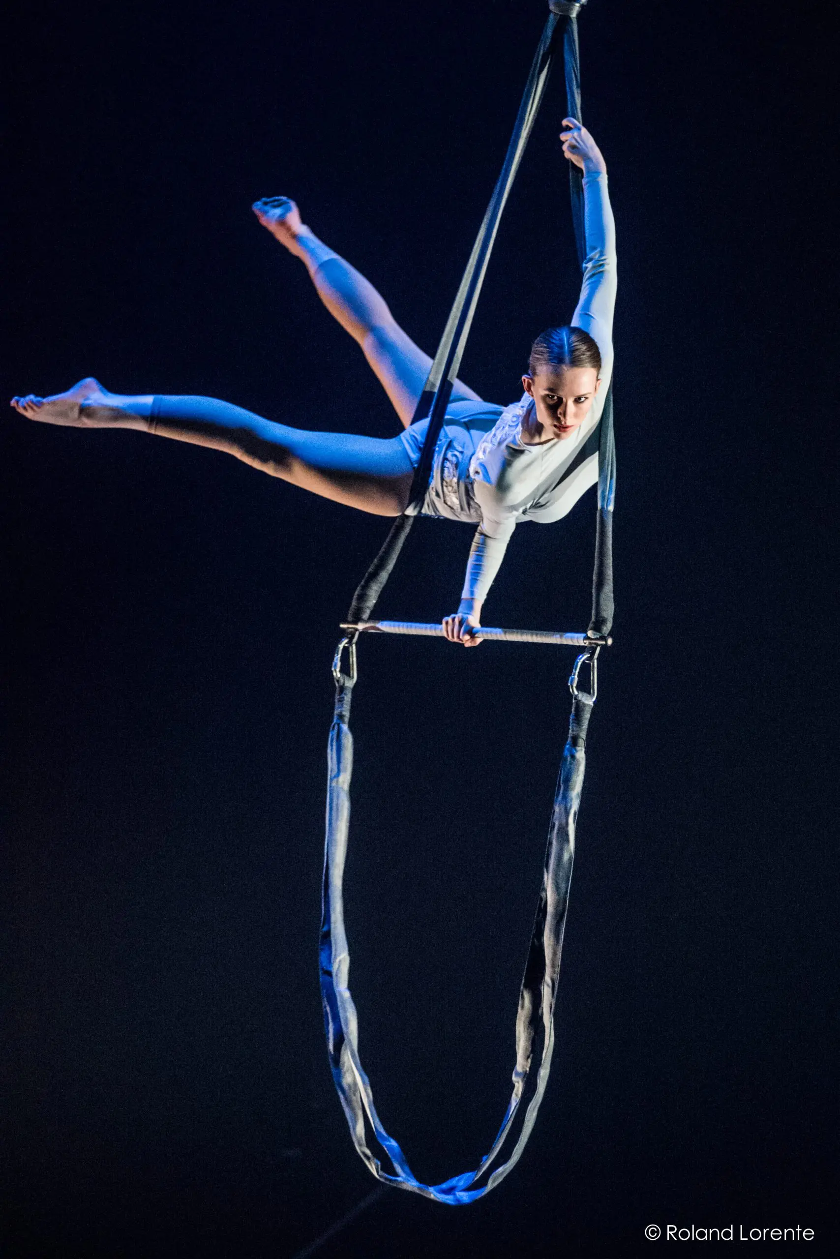 Une artiste aérienne, Halle Baart, vêtue d'un costume blanc, exécute une pose acrobatique sur un appareil suspendu ressemblant à un trapèze, se tenant en équilibre sur un bras tout en étendant gracieusement ses jambes sur un fond sombre.