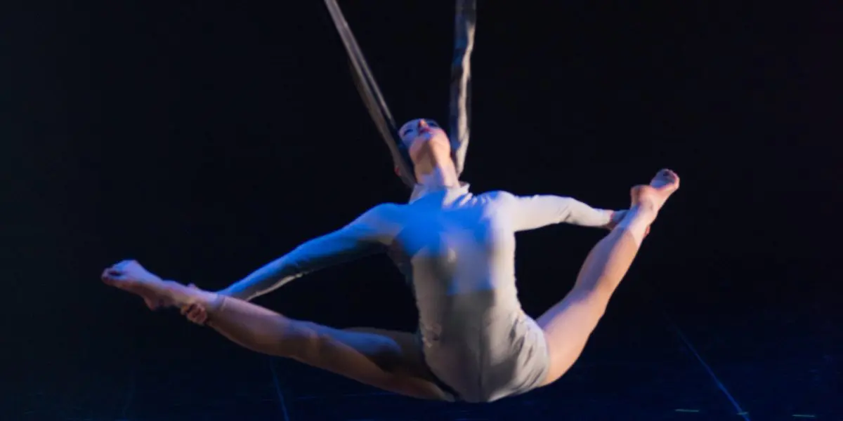 L'artiste aérienne Halle Baart, vêtue d'un justaucorps blanc, effectue un grand écart en étant suspendue à des sangles au-dessus d'une scène sombre, éclairée par des projecteurs spectaculaires.