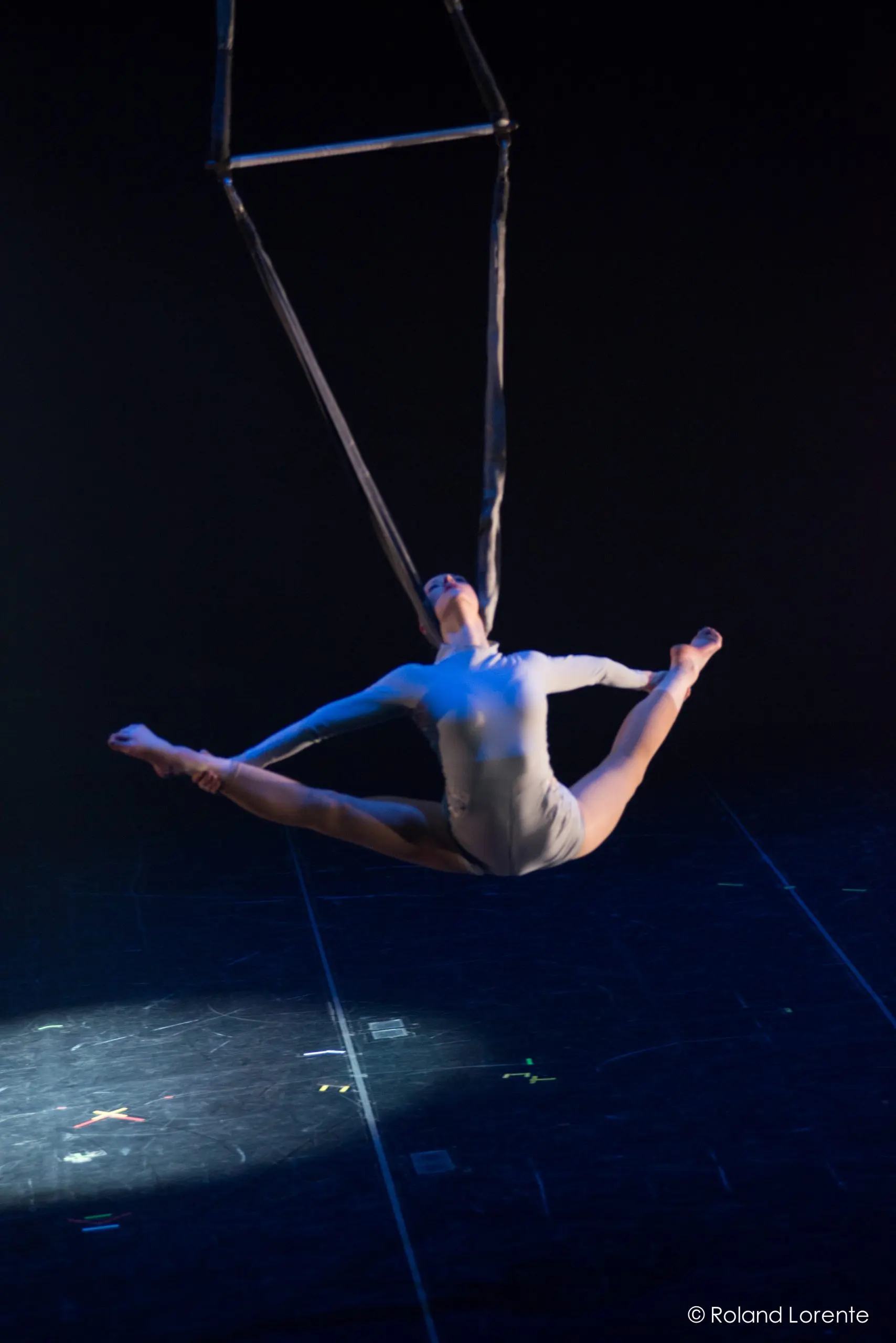 L'artiste aérienne Halle Baart, vêtue d'un justaucorps blanc, effectue un grand écart en étant suspendue à des sangles au-dessus d'une scène sombre, éclairée par des projecteurs spectaculaires.