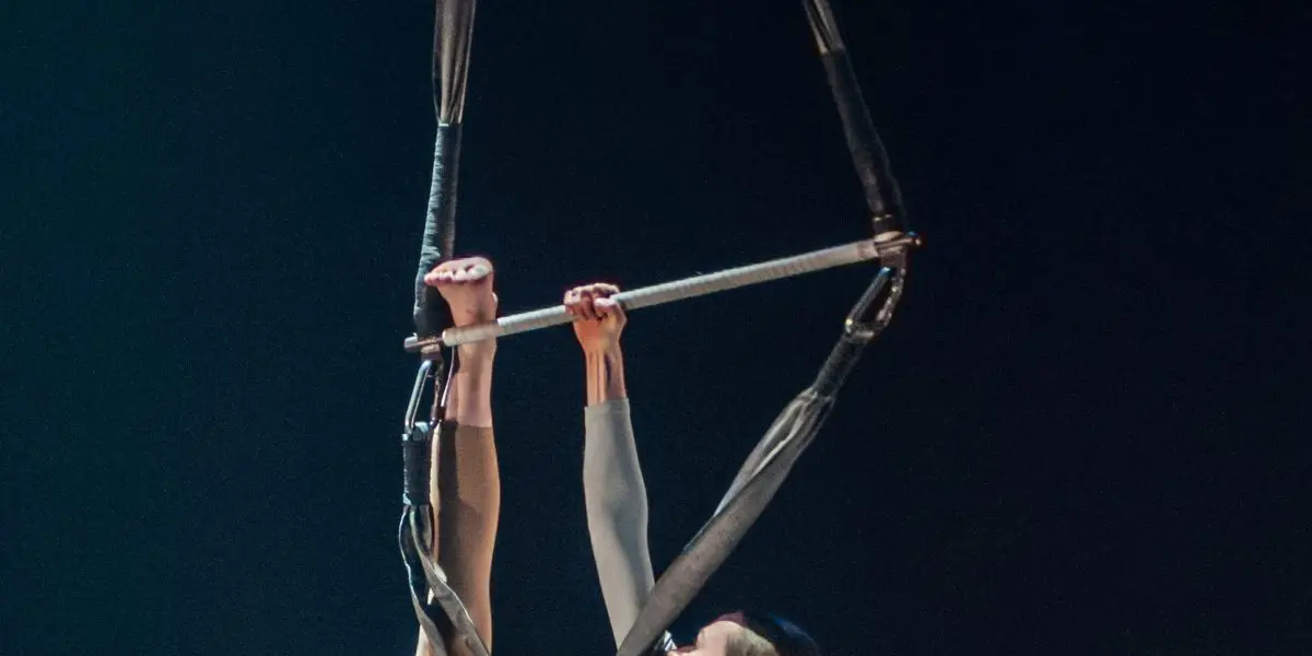 Une artiste aérienne, Halle Baart, exécute une impressionnante pose en deux temps en se suspendant à des sangles et à un cerceau, en se tenant en équilibre avec ses pieds et en saisissant l'appareil d'une main sur un fond sombre.