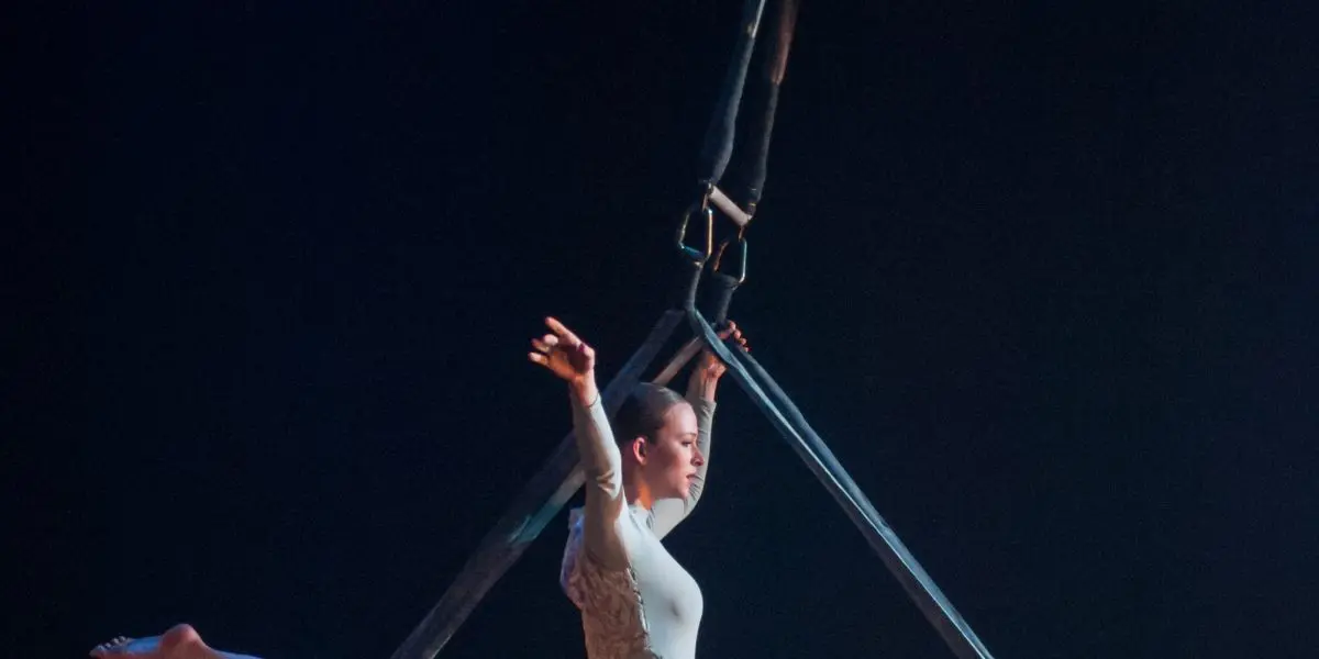 Une femme vêtue d'un justaucorps blanc exécute une fente sur des sangles aériennes, suspendue dans les airs sur un fond sombre, avec un éclairage de scène dramatique mettant en valeur sa pose - une démonstration impressionnante de force et de grâce de la part de Halle Baart.