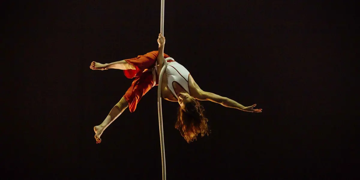 L'artiste aérienne Saffi Watson, vêtue d'un haut blanc et d'un pantalon rouge, est suspendue à l'envers à une corde blanche, les bras et les jambes gracieusement étendus, sur un fond sombre.