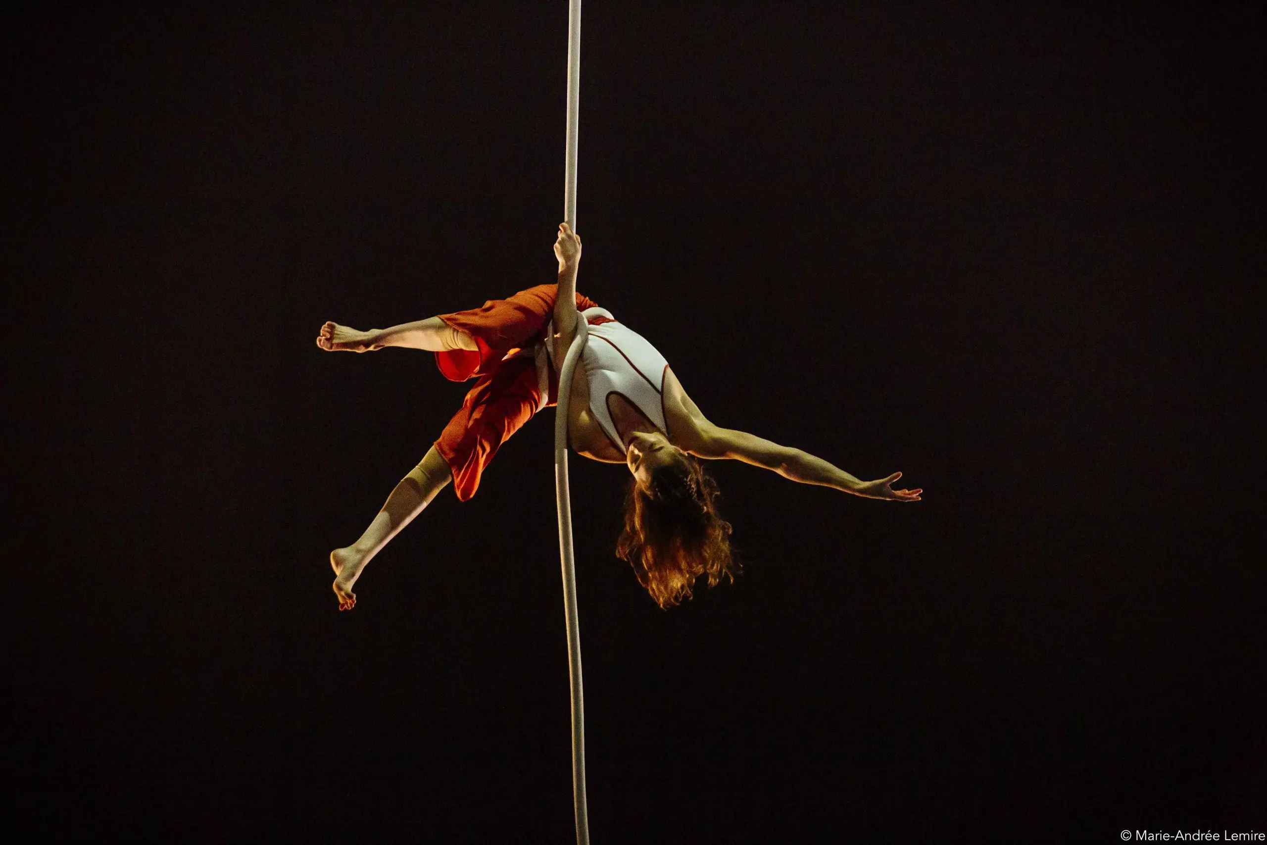 L'artiste aérienne Saffi Watson, vêtue d'un haut blanc et d'un pantalon rouge, est suspendue à l'envers à une corde blanche, les bras et les jambes gracieusement étendus, sur un fond sombre.