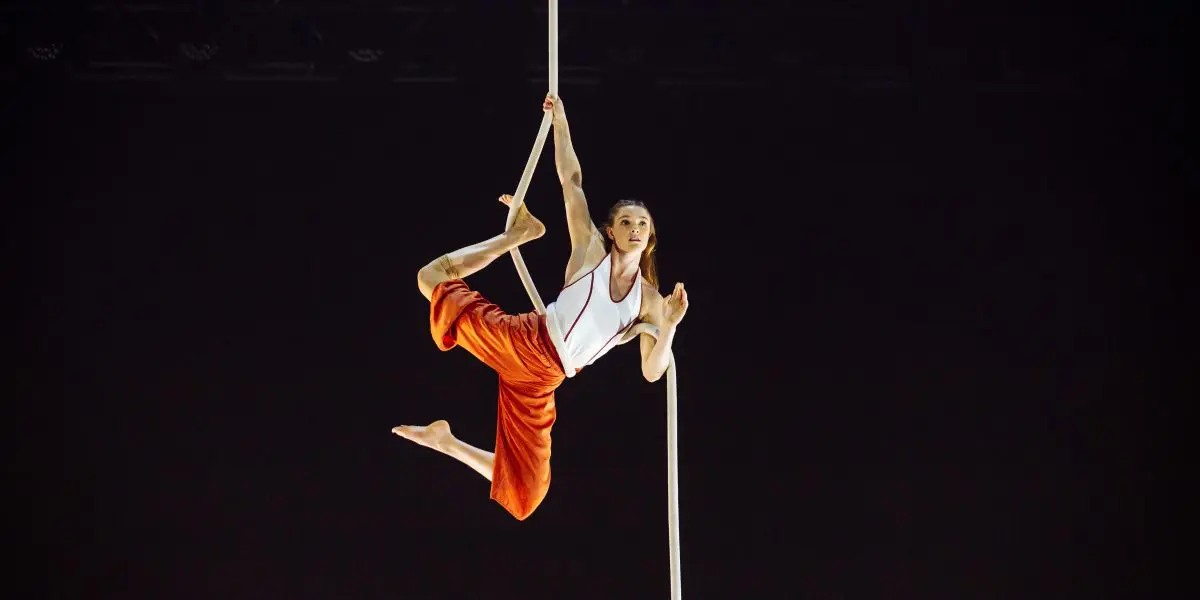 L'artiste aérienne Rosaleen Rogmans, vêtue d'un pantalon orange et d'un haut blanc, pose gracieusement en se suspendant à une corde verticale blanche sur un fond sombre.