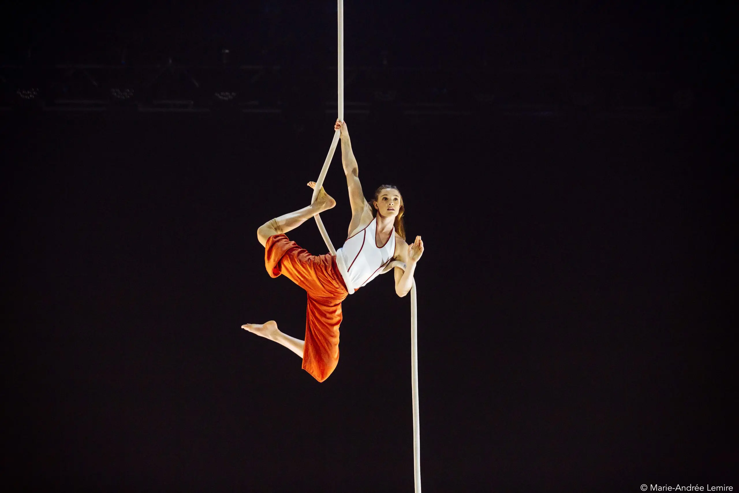 L'artiste aérienne Rosaleen Rogmans, vêtue d'un pantalon orange et d'un haut blanc, pose gracieusement en se suspendant à une corde verticale blanche sur un fond sombre.