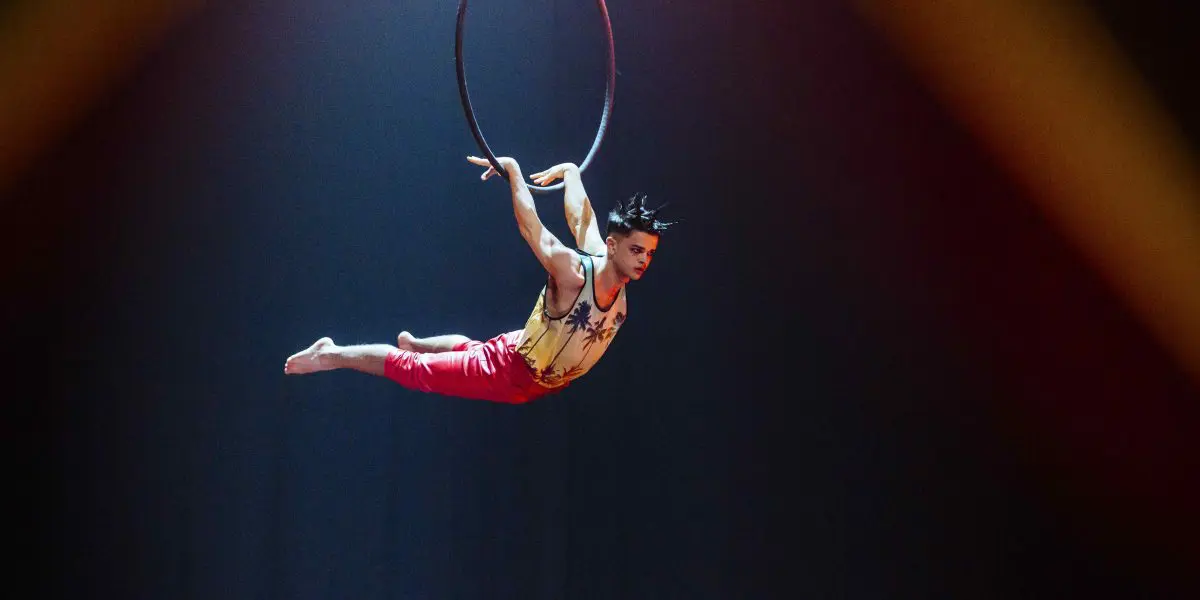 Un artiste de cirque, Enzo Pebre, est suspendu horizontalement à un cerceau aérien, étendant son corps avec les orteils pointus et un bras agrippé au cerceau. Il porte un haut sans manches et un pantalon rouge, avec un éclairage de scène dramatique derrière lui.