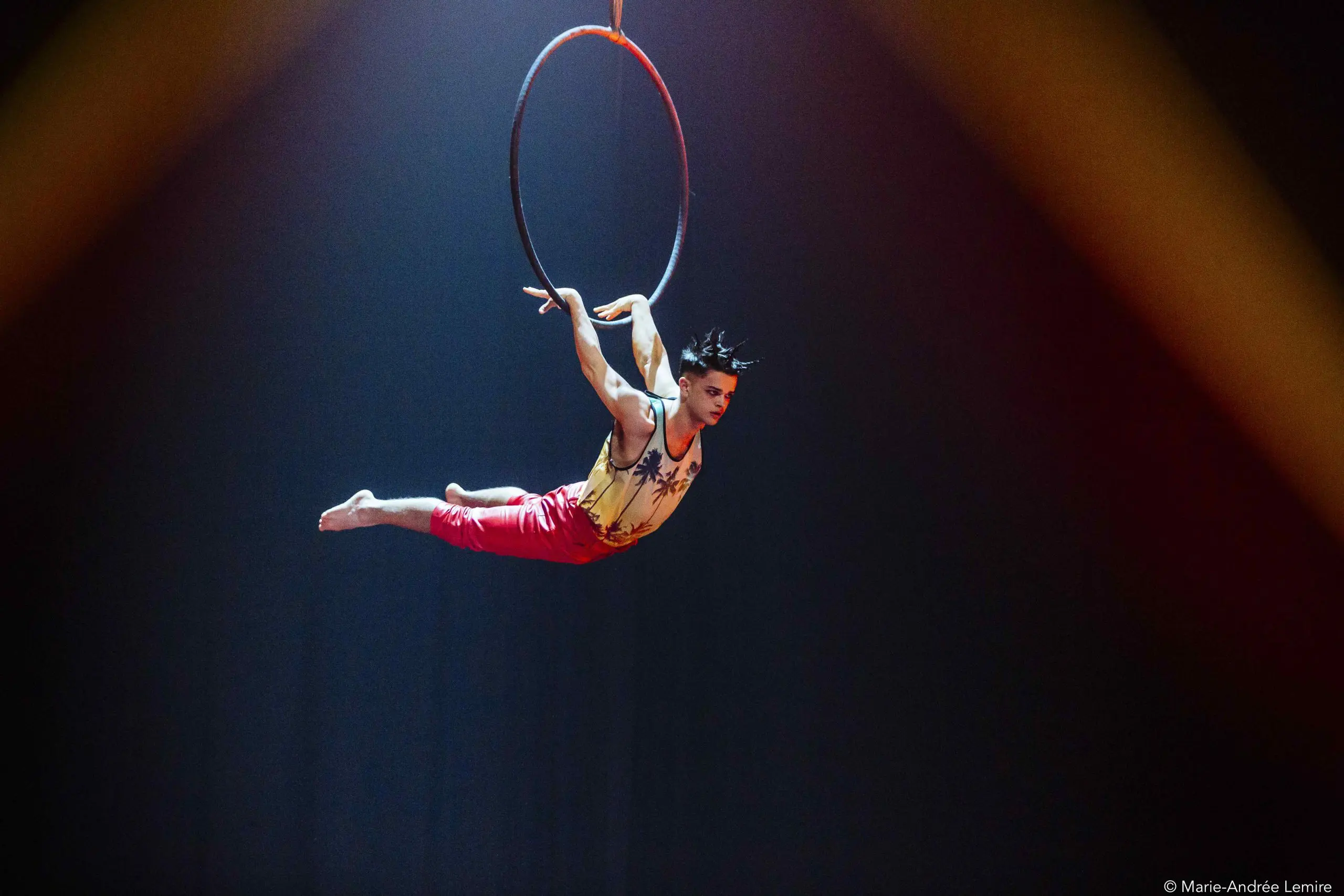 Un artiste de cirque, Enzo Pebre, est suspendu horizontalement à un cerceau aérien, étendant son corps avec les orteils pointus et un bras agrippé au cerceau. Il porte un haut sans manches et un pantalon rouge, avec un éclairage de scène dramatique derrière lui.