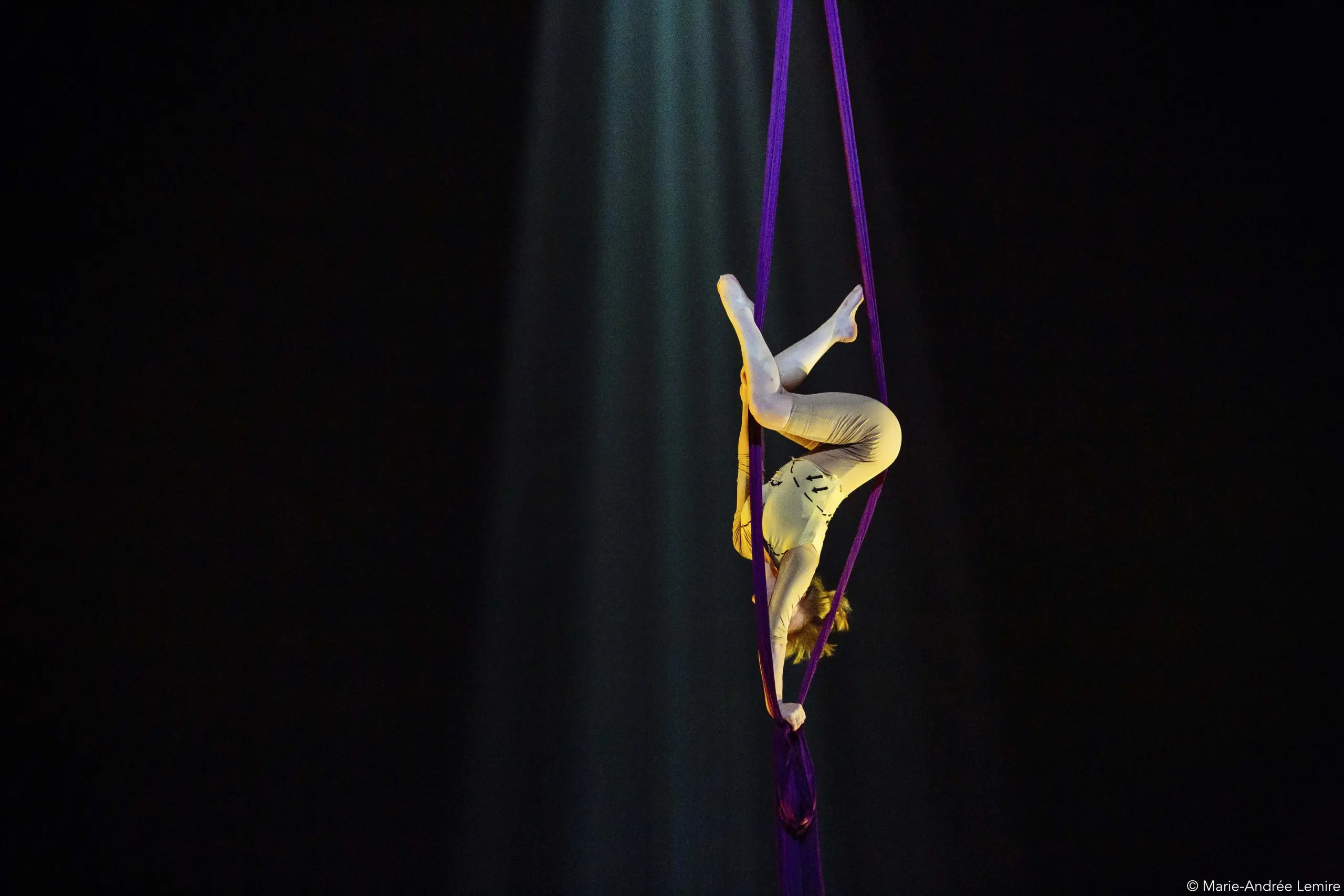L'artiste aérienne Margaux Wasteels, vêtue d'un costume blanc, exécute une pose gracieuse alors qu'elle est suspendue en l'air sur des soies violettes, éclairée par un seul projecteur sur un fond sombre.