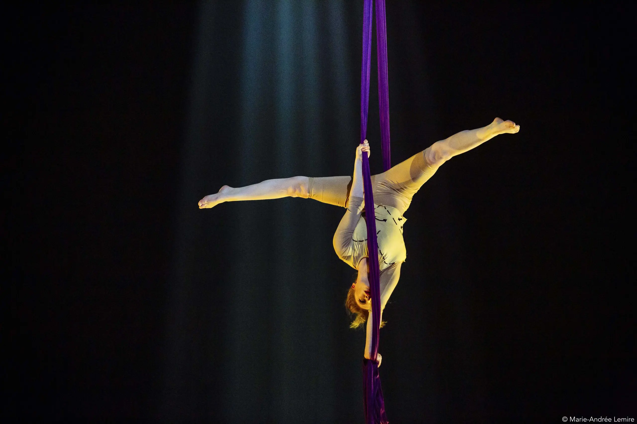Margaux Wasteels, artiste aérienne sur soie, est suspendue à l'envers dans une pose fendue, agrippée à des soies violettes, tandis qu'un projecteur illumine son corps sur un fond sombre.