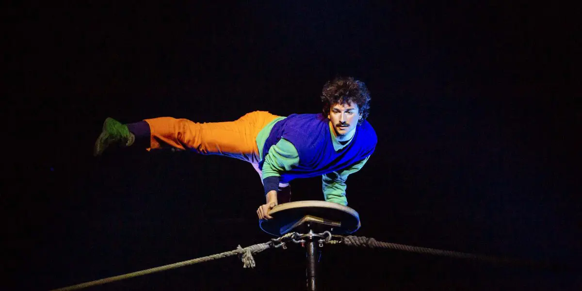 Simon Lemire, vêtu d'un vêtement coloré, se tient en équilibre horizontal sur une main au sommet d'une plate-forme circulaire fixée par deux cordes, sur un fond sombre.
