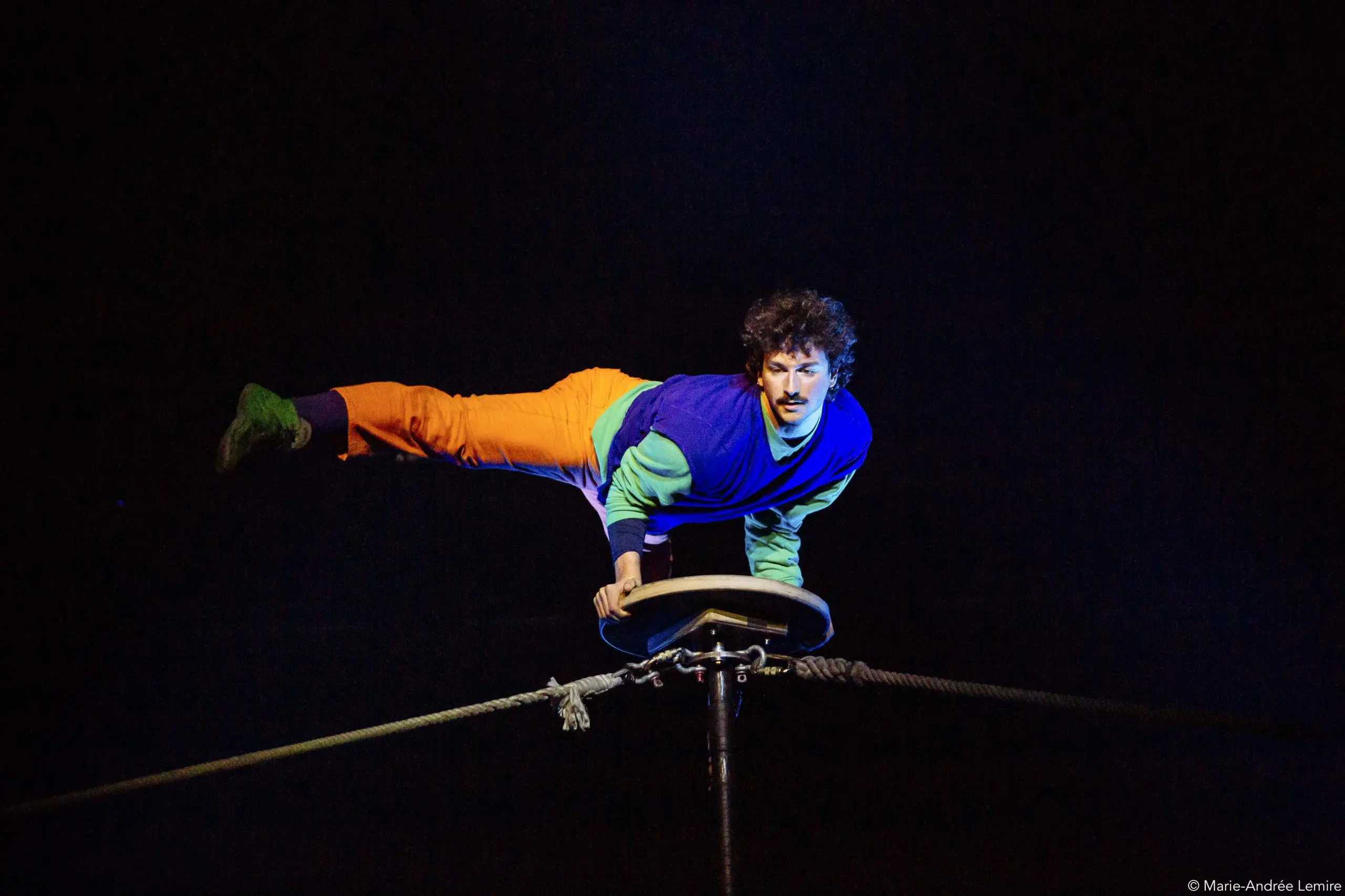 Simon Lemire, vêtu d'un vêtement coloré, se tient en équilibre horizontal sur une main au sommet d'une plate-forme circulaire fixée par deux cordes, sur un fond sombre.