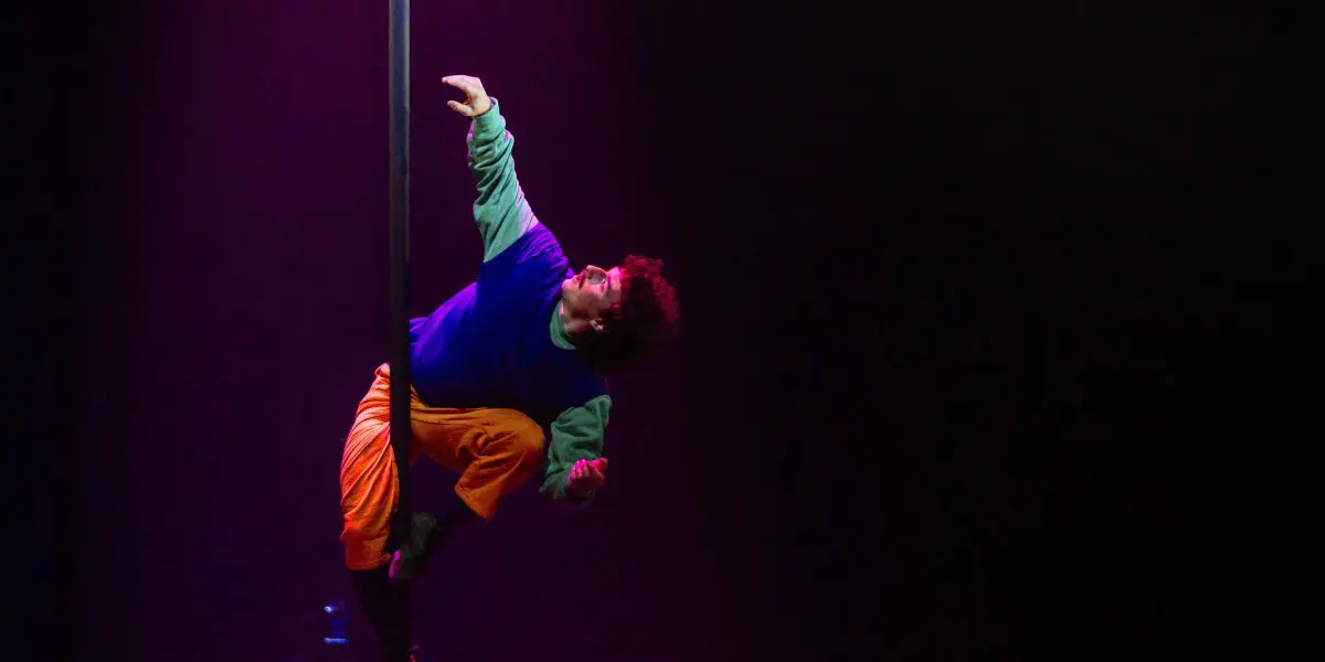 L'artiste Simon Lemire, vêtu de vêtements colorés, grimpe à un poteau vertical sur une scène sombre baignée d'un éclairage violet dramatique.