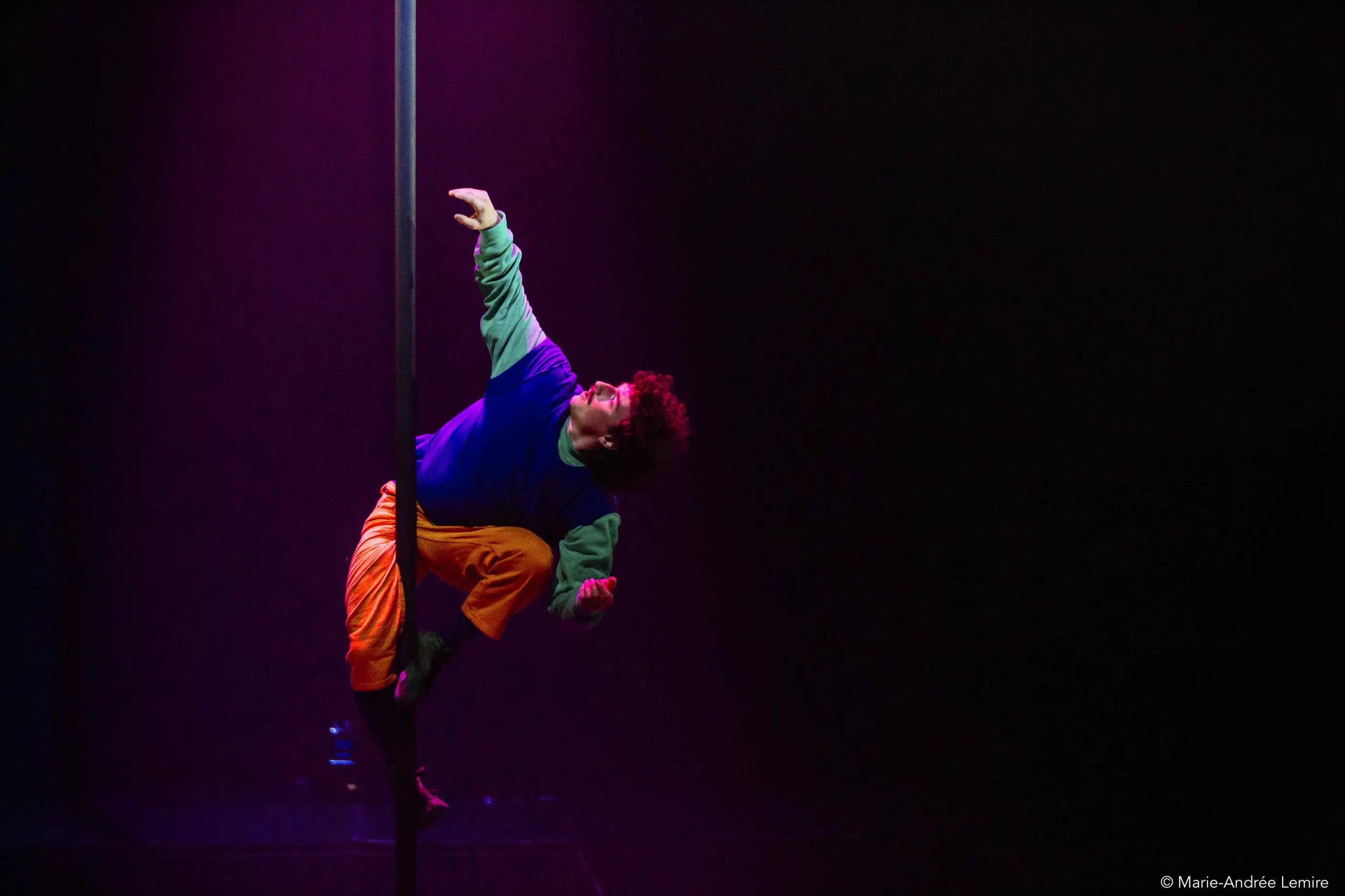 L'artiste Simon Lemire, vêtu de vêtements colorés, grimpe à un poteau vertical sur une scène sombre baignée d'un éclairage violet dramatique.