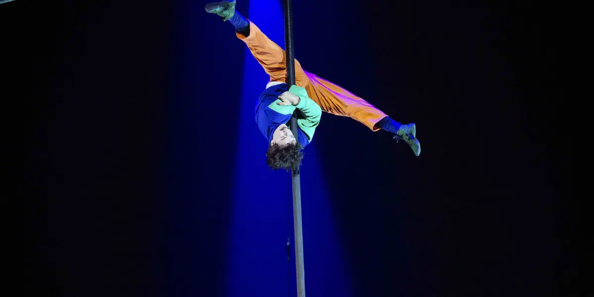 Un artiste de cirque, Simon Lemire, vêtu d'un pantalon orange et d'un haut bleu, est suspendu à l'envers à un poteau vertical sous un projecteur, les jambes écartées et les bras agrippés au poteau, sur un fond sombre.