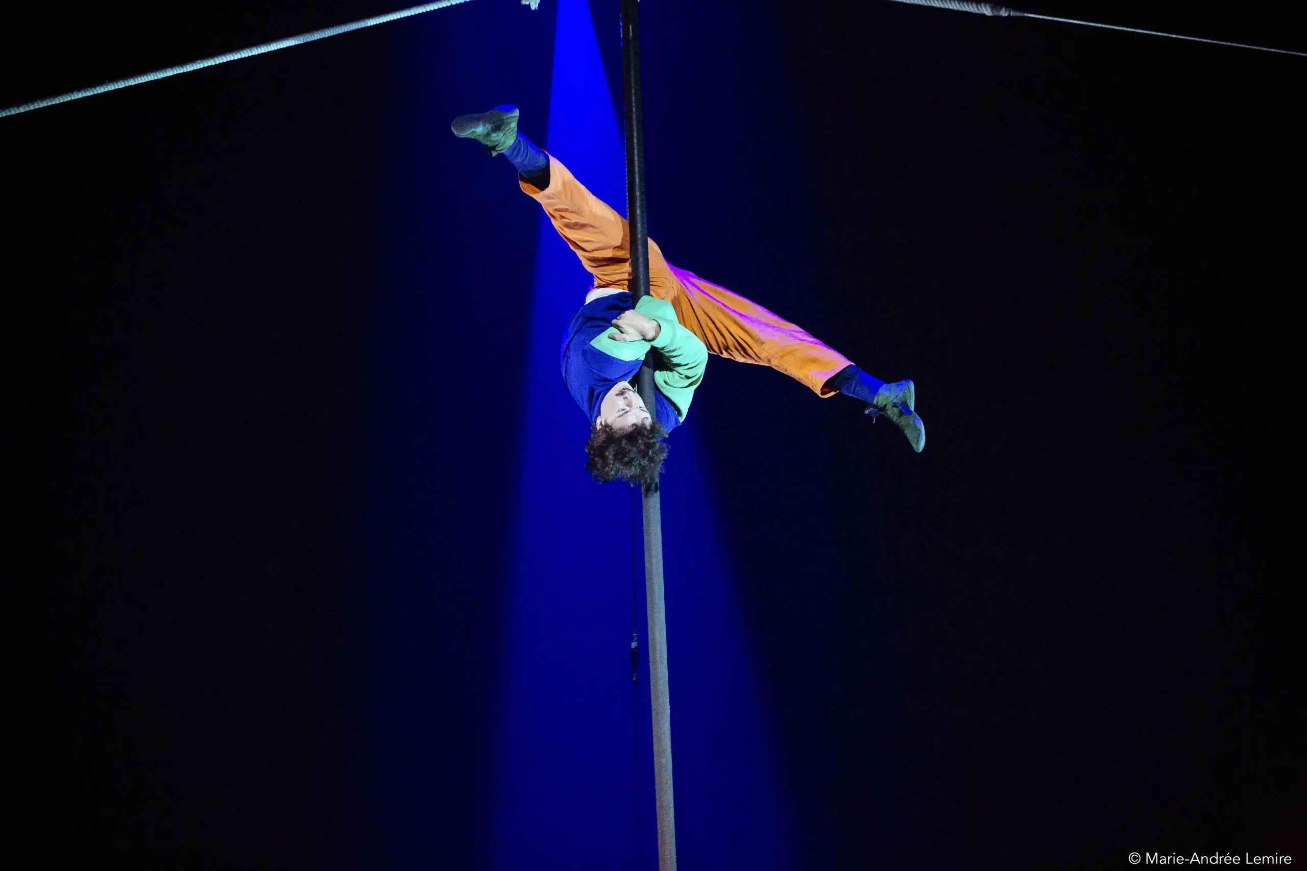 Un artiste de cirque, Simon Lemire, vêtu d'un pantalon orange et d'un haut bleu, est suspendu à l'envers à un poteau vertical sous un projecteur, les jambes écartées et les bras agrippés au poteau, sur un fond sombre.