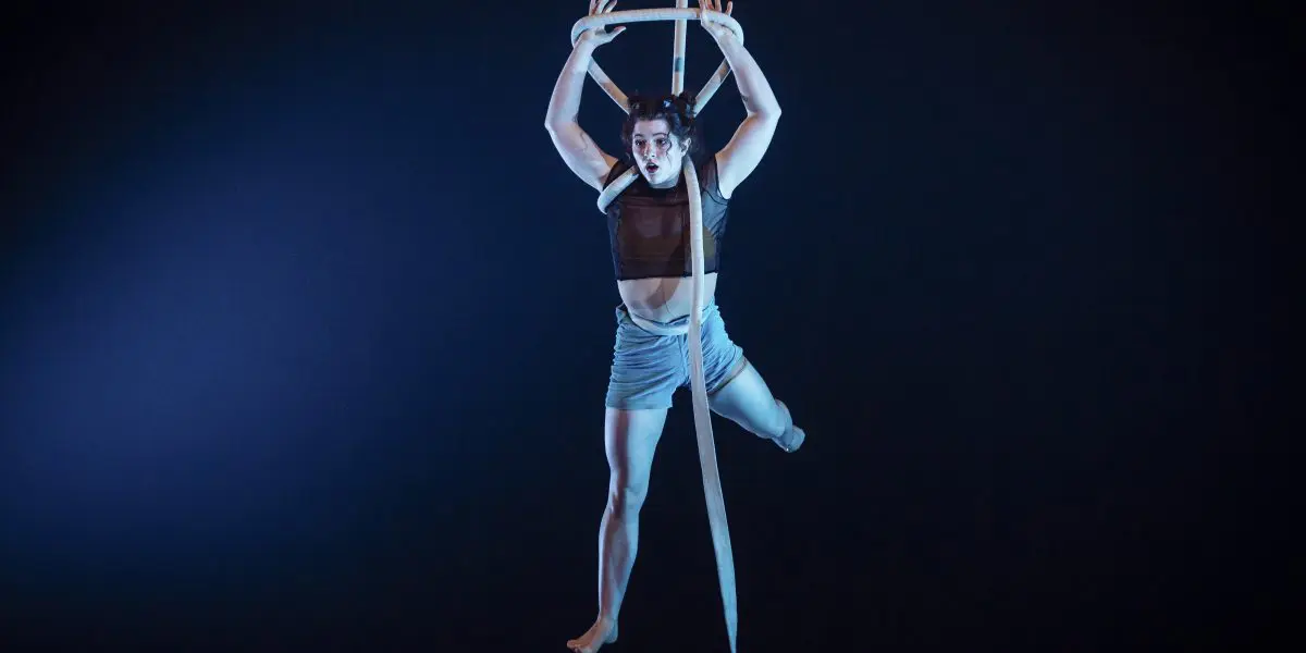 La performeuse aérienne Ellie Rossi est suspendue dans les airs sur des soies blanches sur fond sombre, une main au-dessus de la tête, les jambes étendues, vêtue d'un crop top sombre et d'un short clair, éclairée par la lumière de la scène.