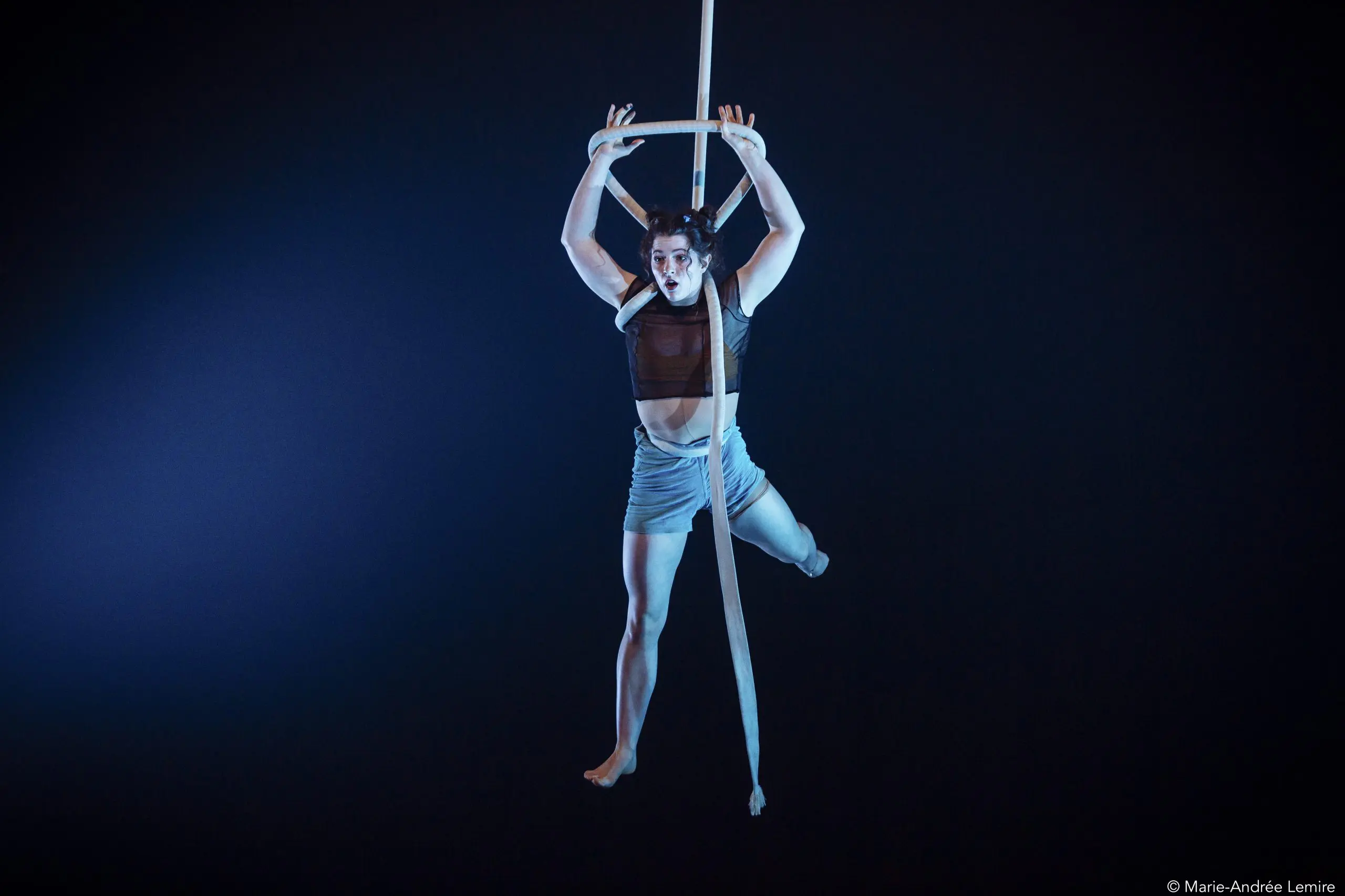 La performeuse aérienne Ellie Rossi est suspendue dans les airs sur des soies blanches sur fond sombre, une main au-dessus de la tête, les jambes étendues, vêtue d'un crop top sombre et d'un short clair, éclairée par la lumière de la scène.