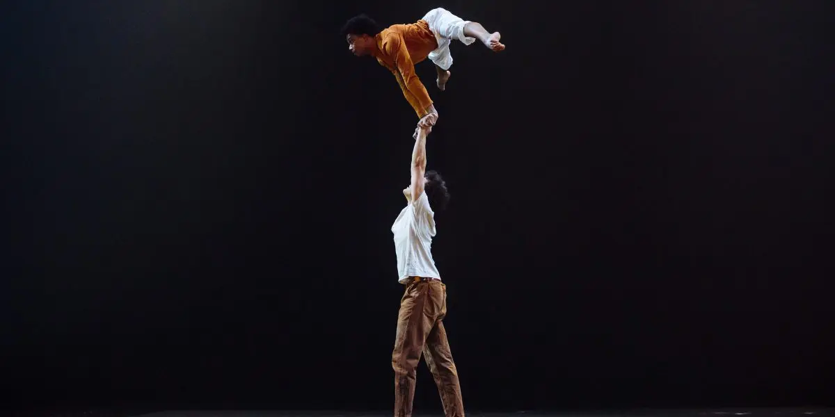 Deux danseurs effectuent un porté acrobatique sur scène ; l'un se tient au sol, en équilibre sur un pied, tandis que l'autre tient l'autre danseur au-dessus de sa tête avec un bras - un moment saisissant dans la chorégraphie d'Oliver Layher sur un fond sombre et vide.