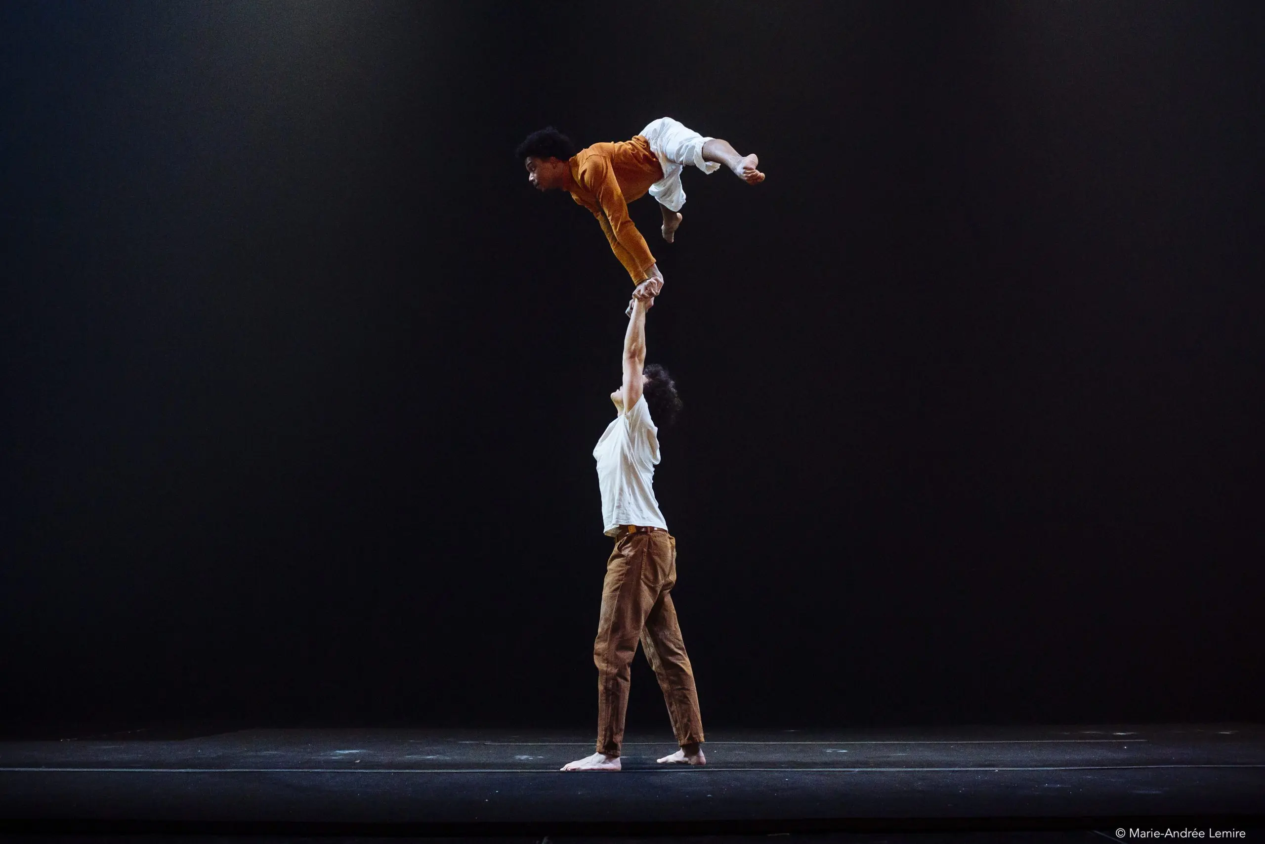 Deux danseurs effectuent un porté acrobatique sur scène ; l'un se tient au sol, en équilibre sur un pied, tandis que l'autre tient l'autre danseur au-dessus de sa tête avec un bras - un moment saisissant dans la chorégraphie d'Oliver Layher sur un fond sombre et vide.