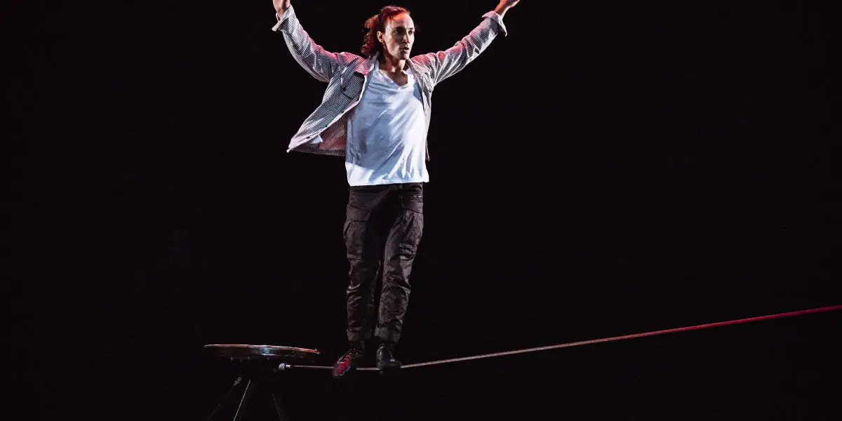 Joel Malkoff, cheveux longs, vêtu d'une chemise blanche et d'une veste légère, se tient en équilibre, les bras levés, tout en marchant sur une corde raide, sur un fond sombre.