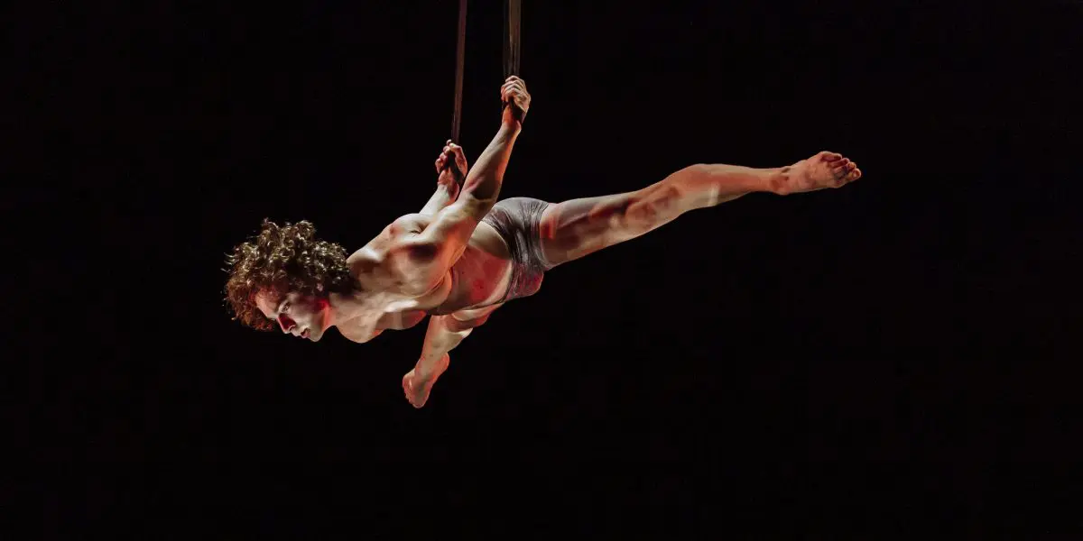 L'acrobate aérien Agustin Rodriguez Beltran s'exécute sur des sangles, suspendu dans les airs avec une jambe tendue, vêtu d'un minimum de vêtements. Un éclairage dramatique met en valeur leur force et leur équilibre sur un fond sombre.