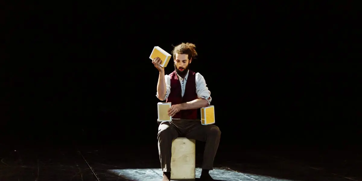 Joaquim Verrier est assis sur une boîte sur scène, jonglant avec trois blocs jaunes sous un projecteur. Vêtu d'un gilet, d'une chemise blanche et d'un pantalon retroussé, il se détache sur le fond sombre qui l'entoure.