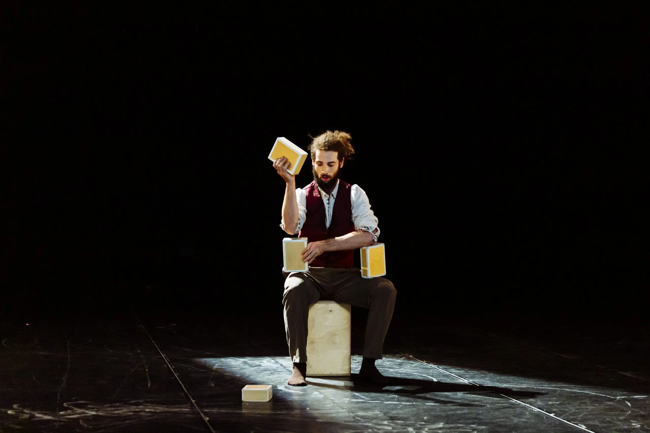 Joaquim Verrier est assis sur une boîte sur scène, jonglant avec trois blocs jaunes sous un projecteur. Vêtu d'un gilet, d'une chemise blanche et d'un pantalon retroussé, il se détache sur le fond sombre qui l'entoure.