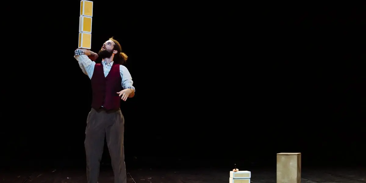 Joaquim Verrier, un artiste en gilet et nœud papillon, tient une pile de boîtes en équilibre sur une main sur une scène sombre, avec une autre boîte et une pile de boîtes sur le sol à proximité.