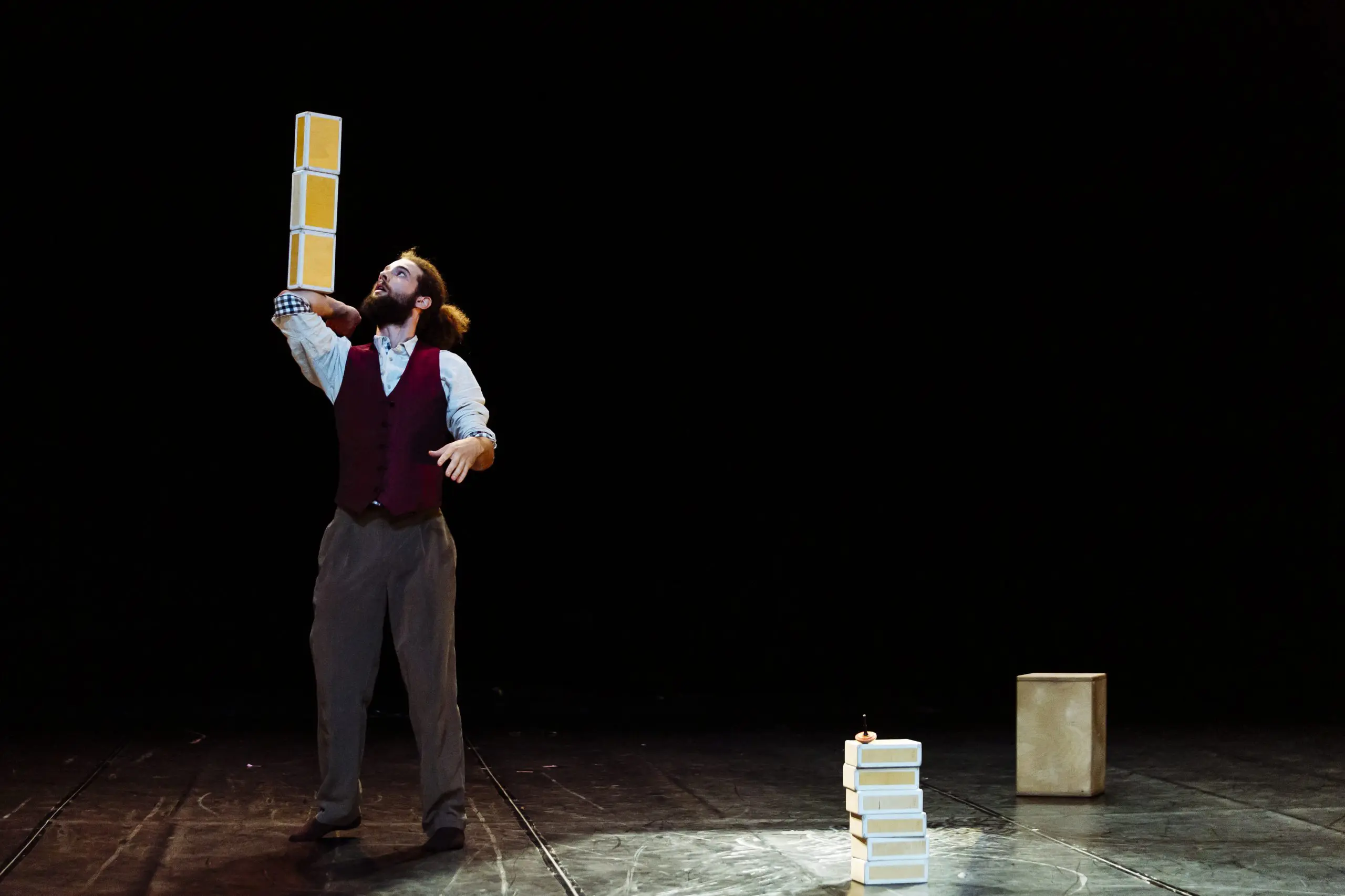 Joaquim Verrier, un artiste en gilet et nœud papillon, tient une pile de boîtes en équilibre sur une main sur une scène sombre, avec une autre boîte et une pile de boîtes sur le sol à proximité.