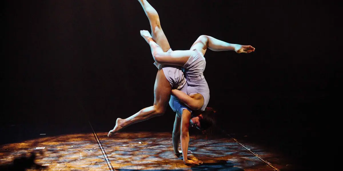 Une interprète, rappelant Léann Gingras et Kaisha Dessalines Wright, se tient en équilibre sur une main dans une tenue bleu clair, prenant une pose acrobatique dynamique sur une scène faiblement éclairée par des lumières dramatiques.
