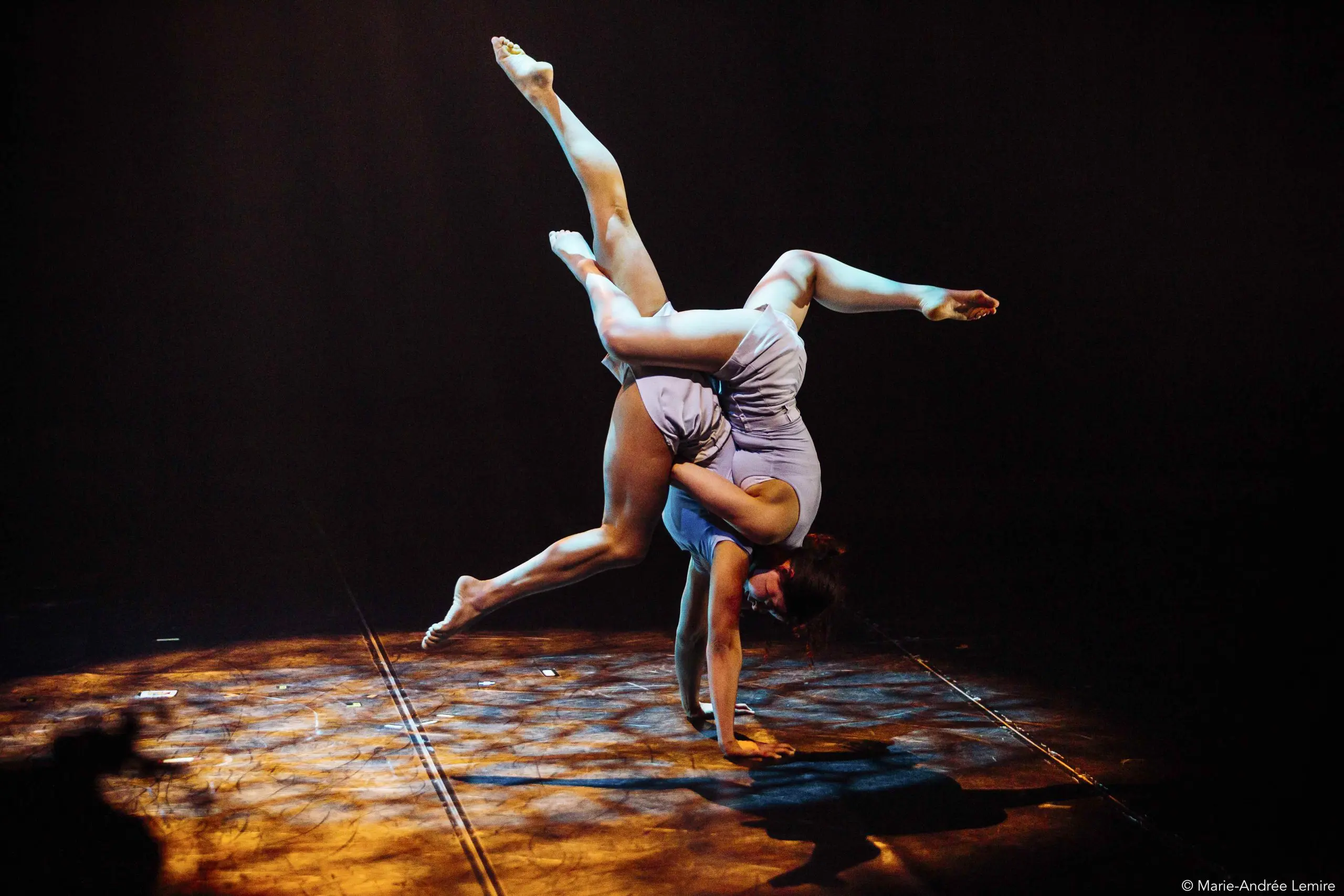 Une interprète, rappelant Léann Gingras et Kaisha Dessalines Wright, se tient en équilibre sur une main dans une tenue bleu clair, prenant une pose acrobatique dynamique sur une scène faiblement éclairée par des lumières dramatiques.