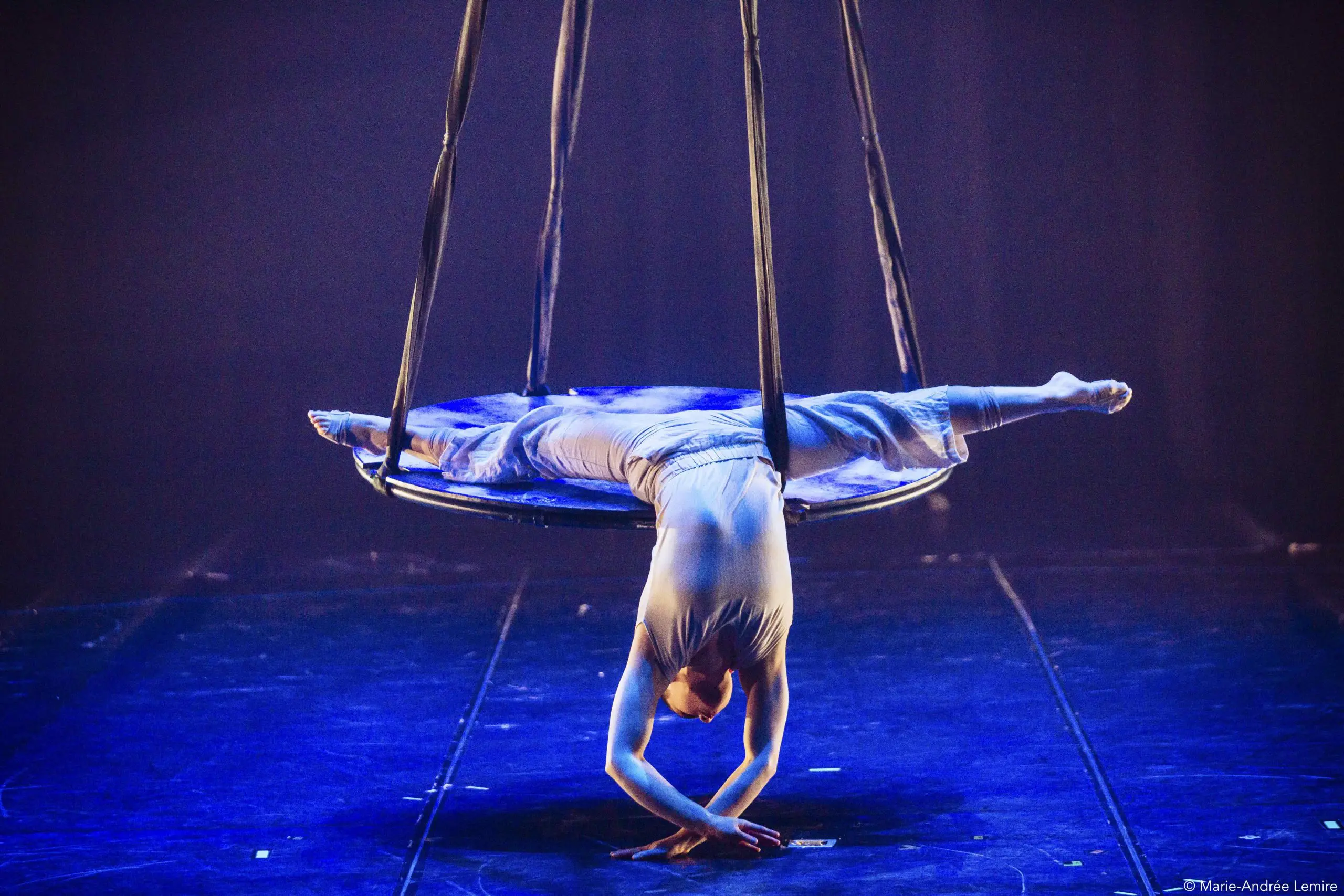 Saffi Watson, une artiste de cirque vêtue d'un costume blanc, est suspendue la tête en bas à un cerceau, en équilibre sur ses mains, les jambes étendues en éventail, sous un éclairage de scène spectaculaire.
