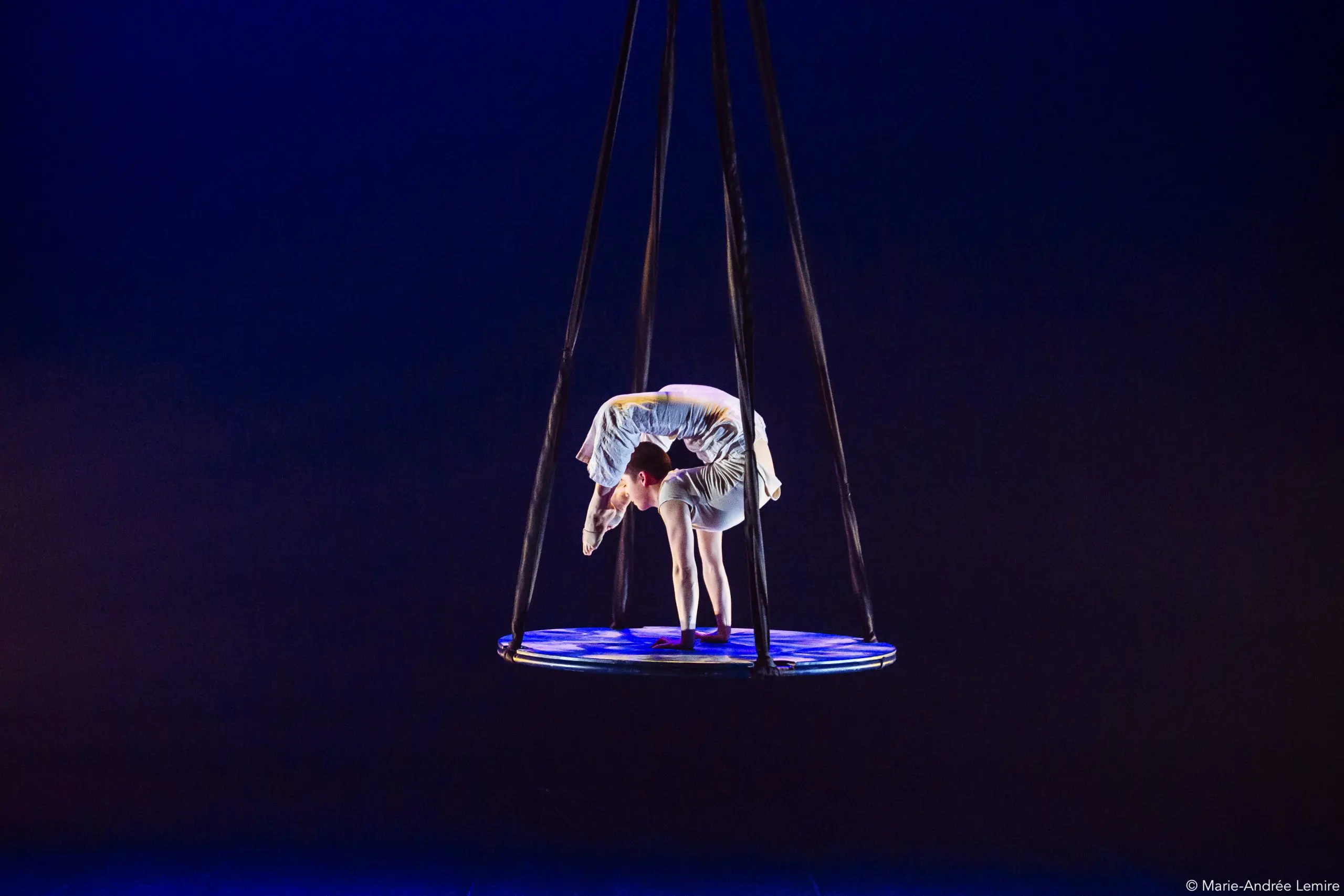 Saffi Watson, artiste de cirque en costume blanc, se tient en équilibre sur un cerceau aérien circulaire, suspendu par des sangles noires, sur une scène sombre à l'éclairage dramatique.