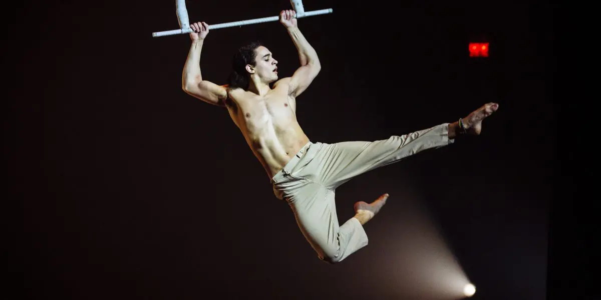 Un acrobate masculin, Zebulon Simoneau, torse nu et vêtu d'un pantalon beige, exécute un mouvement aérien dynamique sur un trapèze sur un fond sombre, avec une jambe tendue et les deux bras agrippés à la barre.