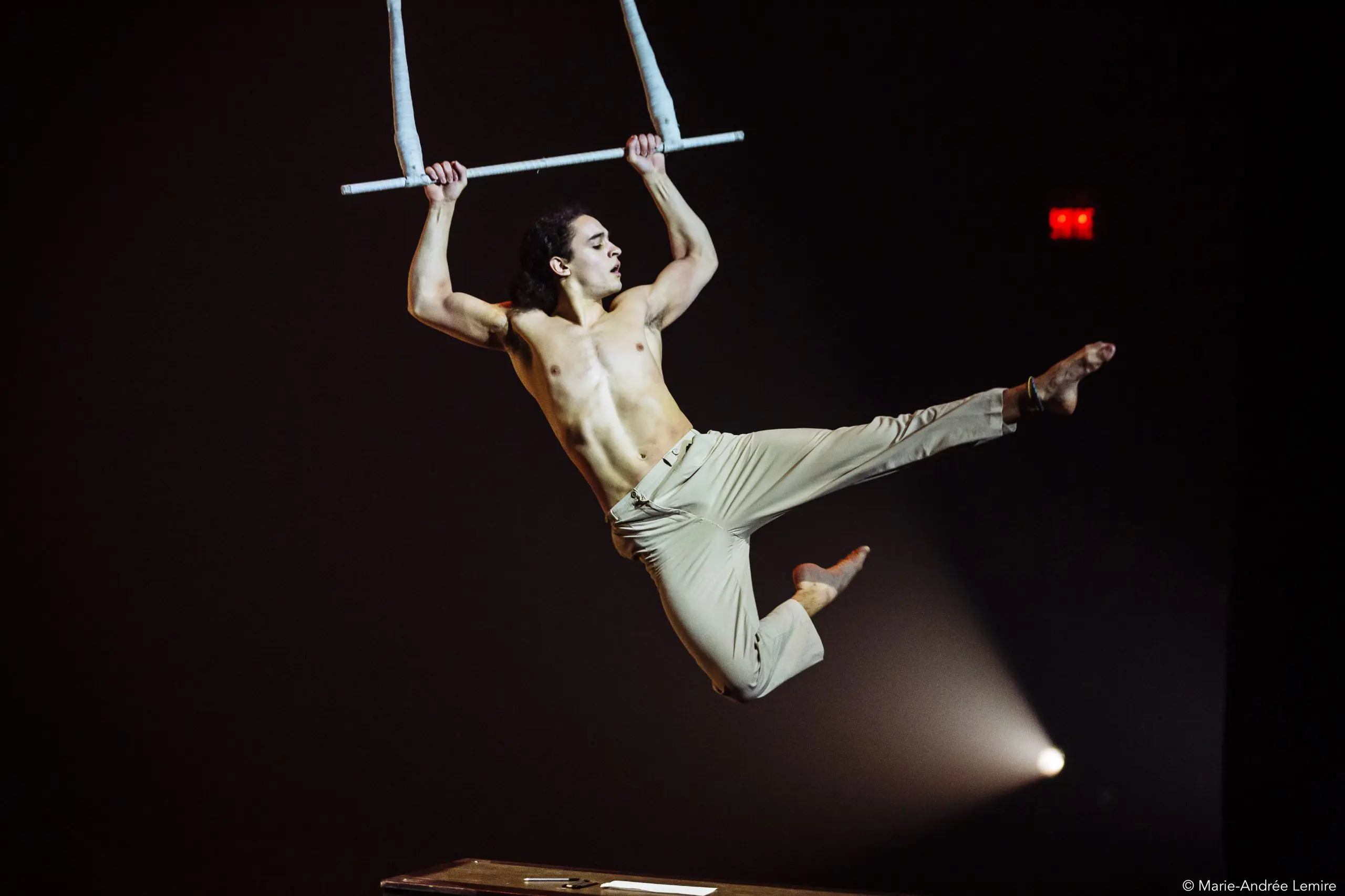Un acrobate masculin, Zebulon Simoneau, torse nu et vêtu d'un pantalon beige, exécute un mouvement aérien dynamique sur un trapèze sur un fond sombre, avec une jambe tendue et les deux bras agrippés à la barre.